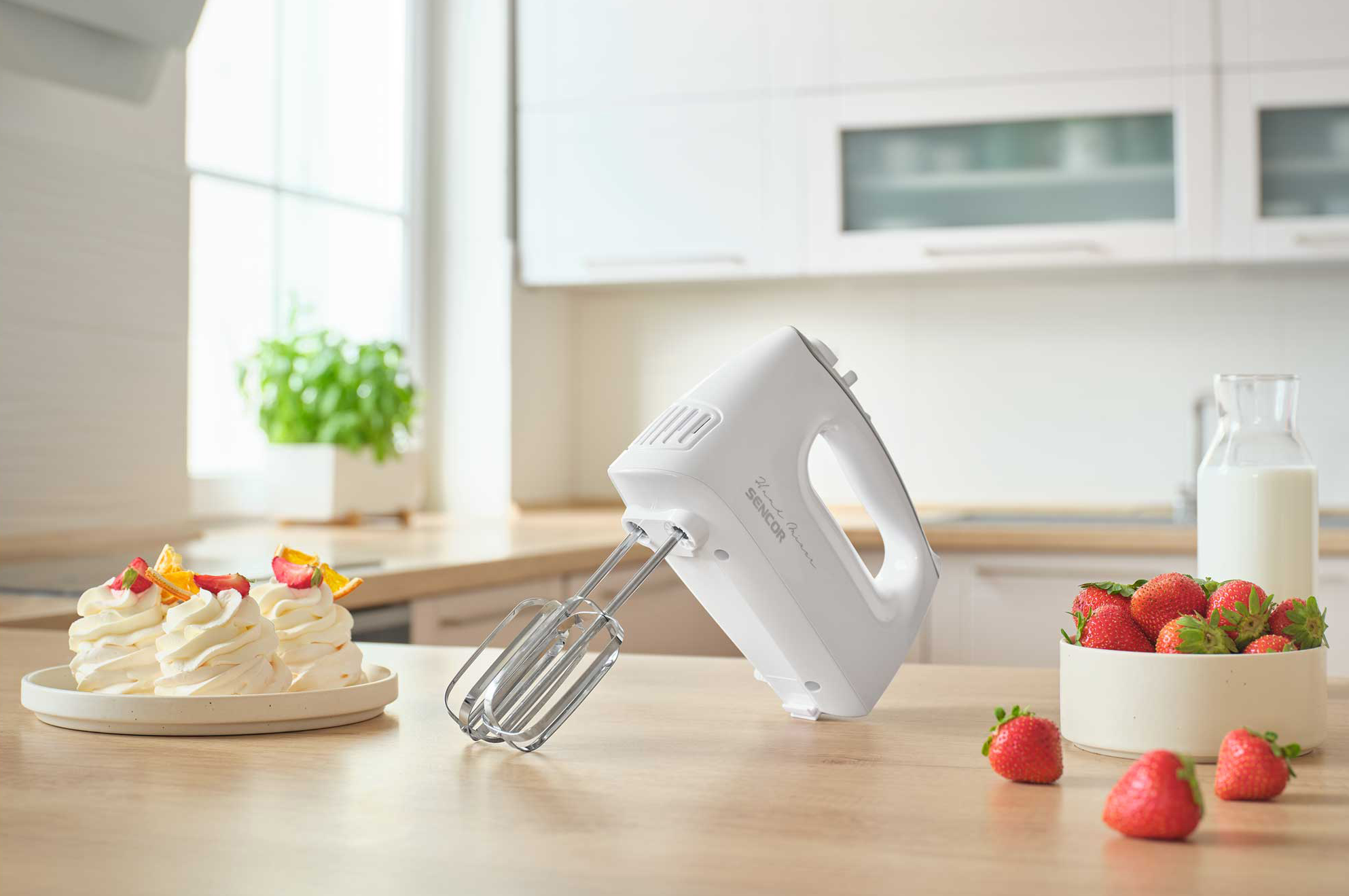 Egy fehér kézi mixer eperrel és süteményekkel egy fali asztalon.