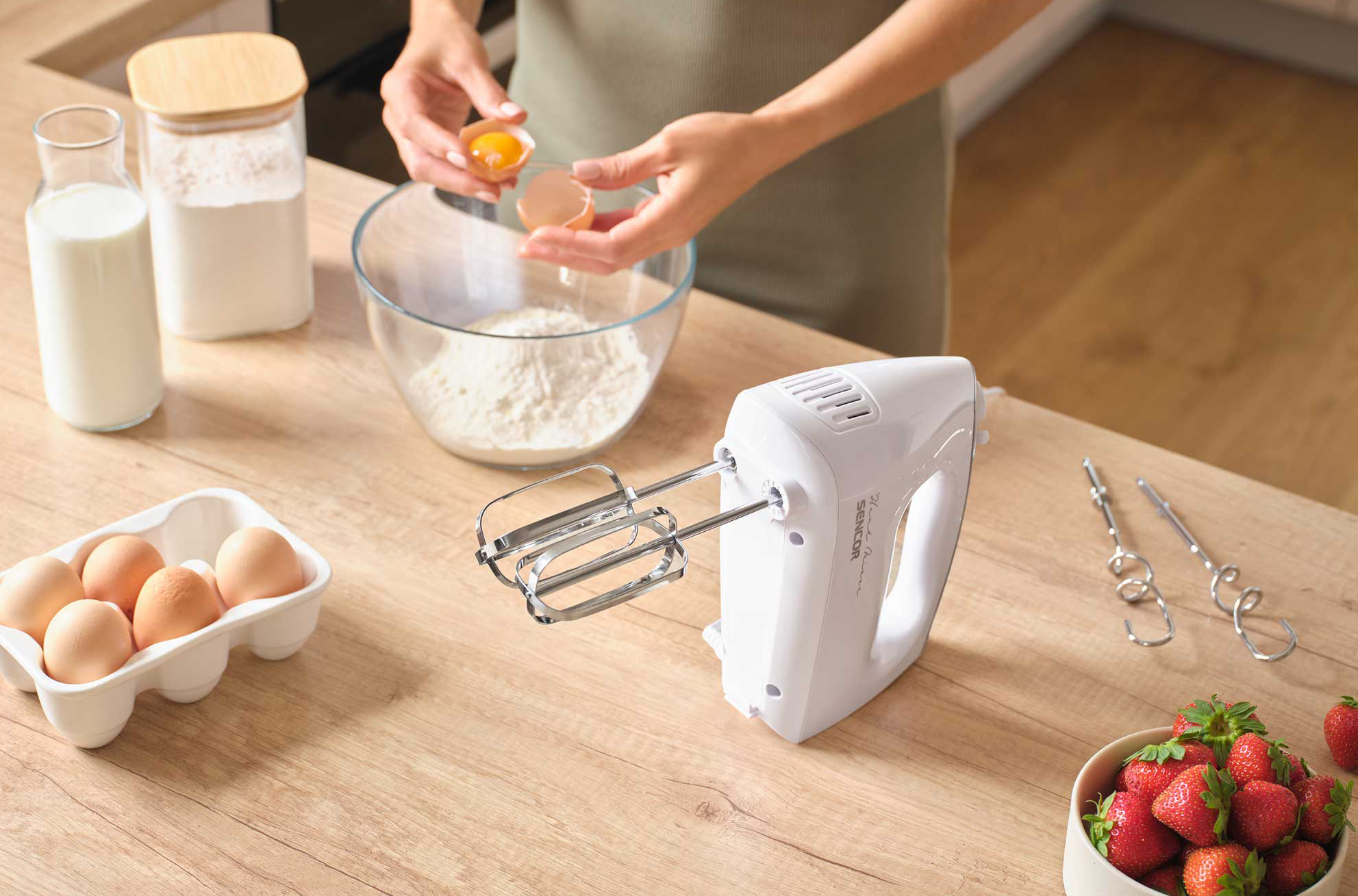 Egy személy tojásokat tör egy lisztes tál fölé, kézi mixer és hozzávalók a közelben.