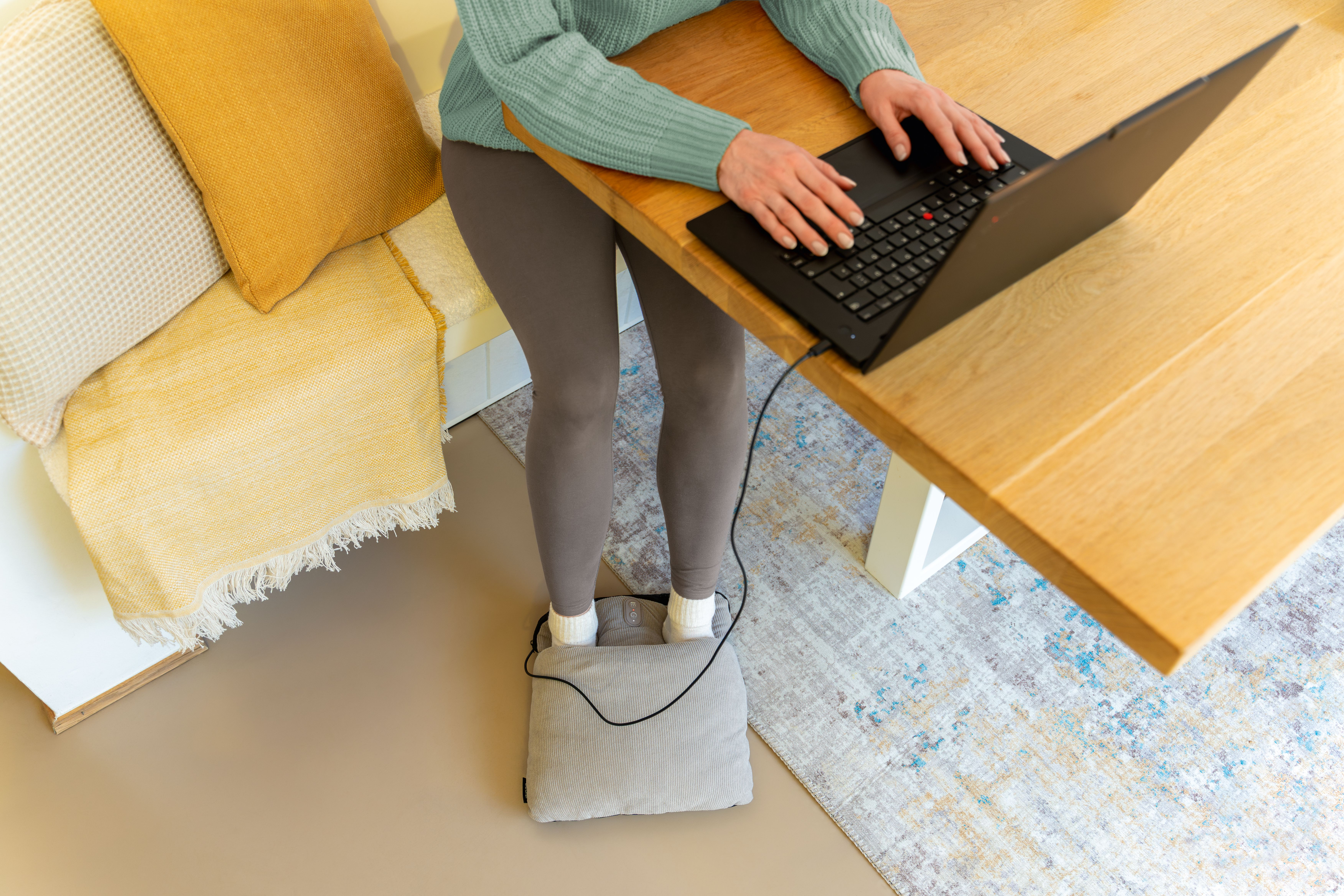 Eine Person steht auf einem Fußwärmer vor einem Holztisch und benutzt einen Laptop.