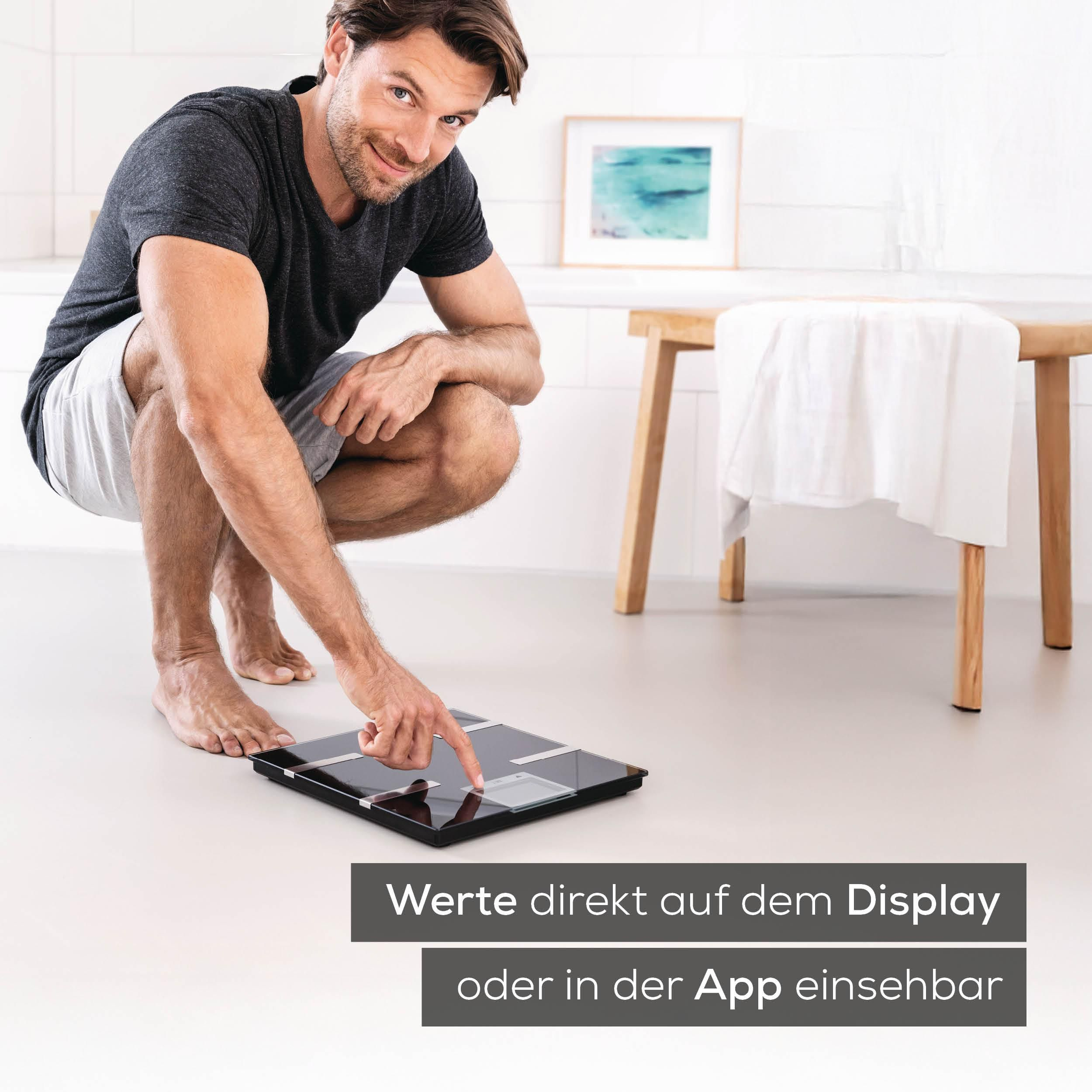 Mann kniet auf einer Waage, die auf dem Boden steht. Er zeigt auf das Display.