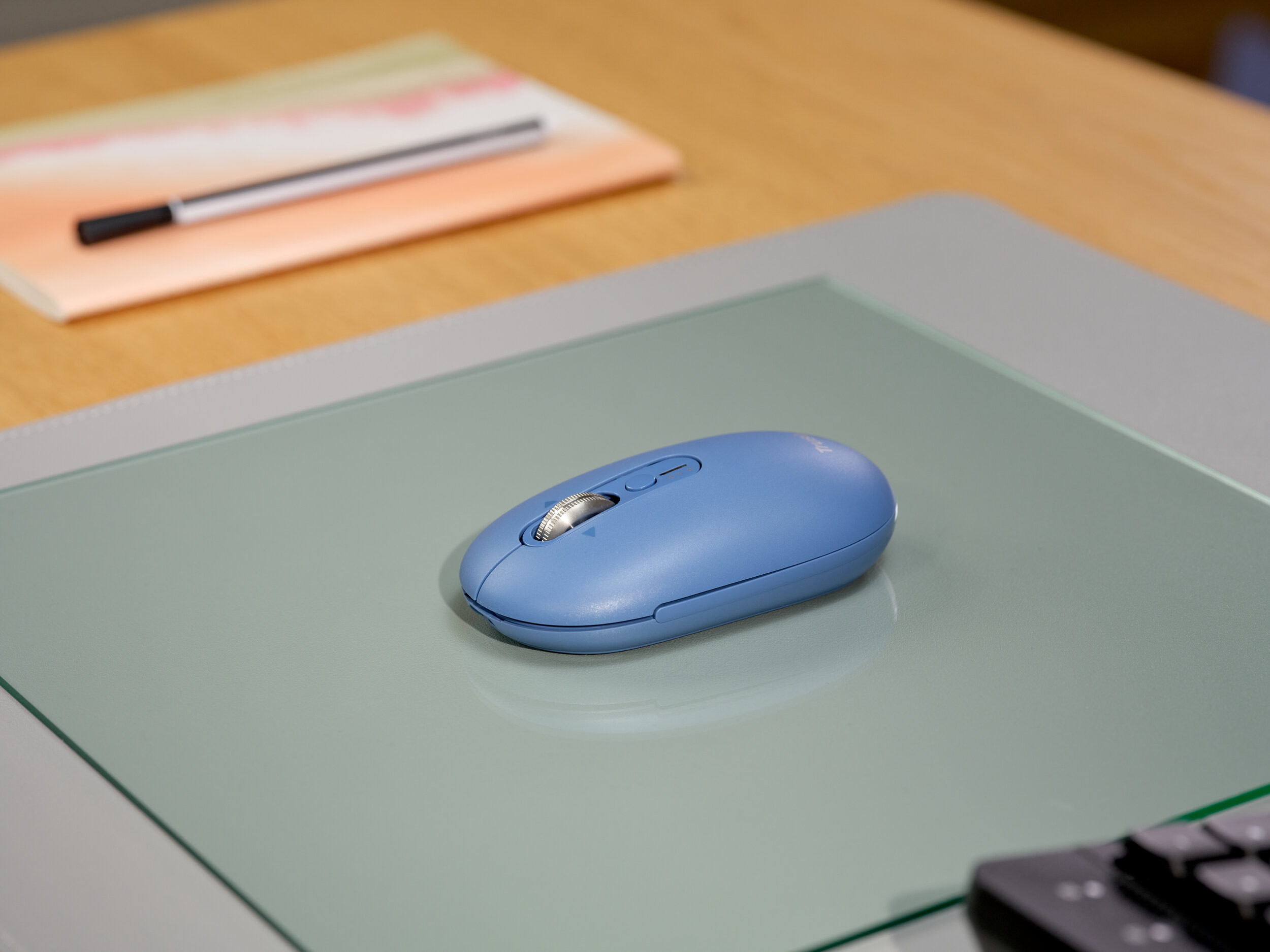 Un mouse wireless blu poggia su un tappetino di vetro accanto a un taccuino e una penna.