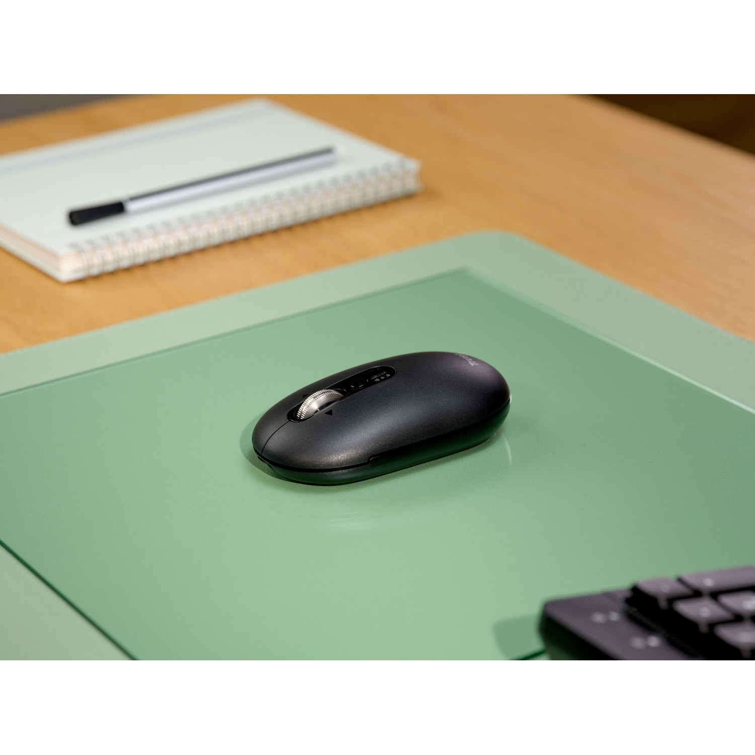 Un mouse scuro poggia su un tappetino verde. Quaderno e penna sullo sfondo.
