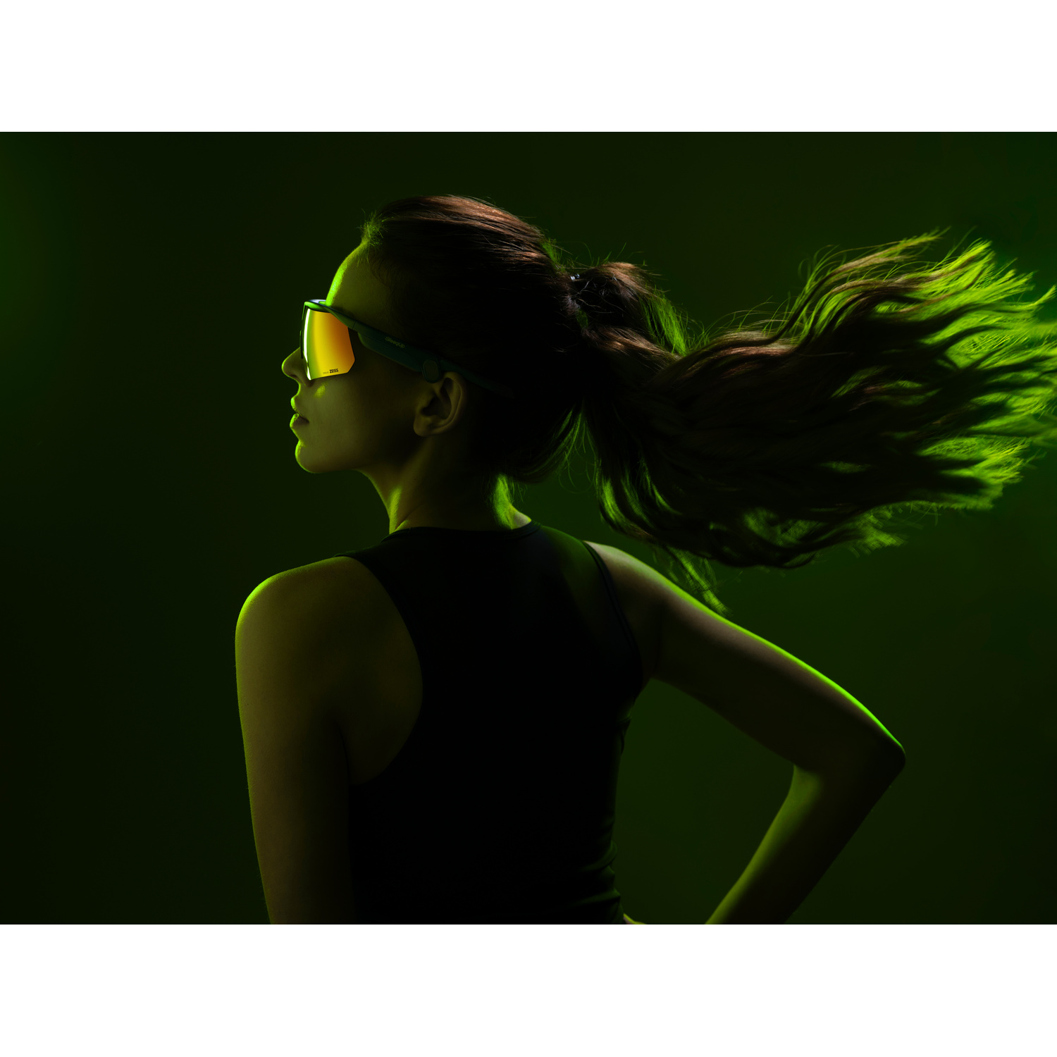 Donna con occhiali sportivi di profilo, capelli che fluttuano nella luce verde.