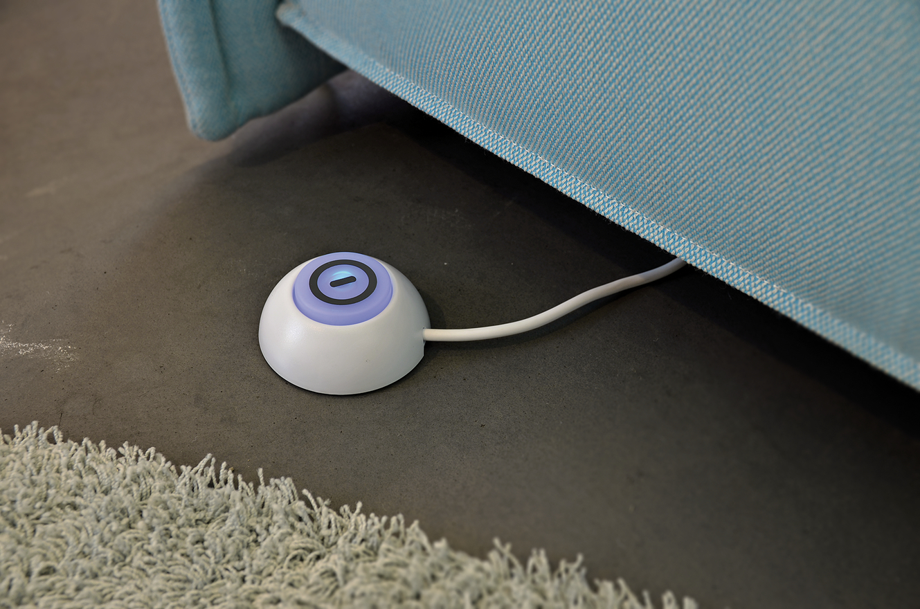 Ein weißer runder Sensor mit blauem Licht liegt auf einem grauen Boden neben einem türkisfarbenen Sofa.