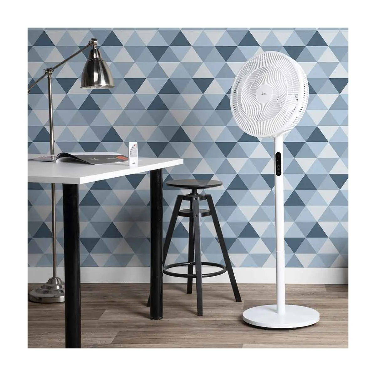 Un ventilador de pie blanco junto a una mesa blanca con lámpara y taburete.