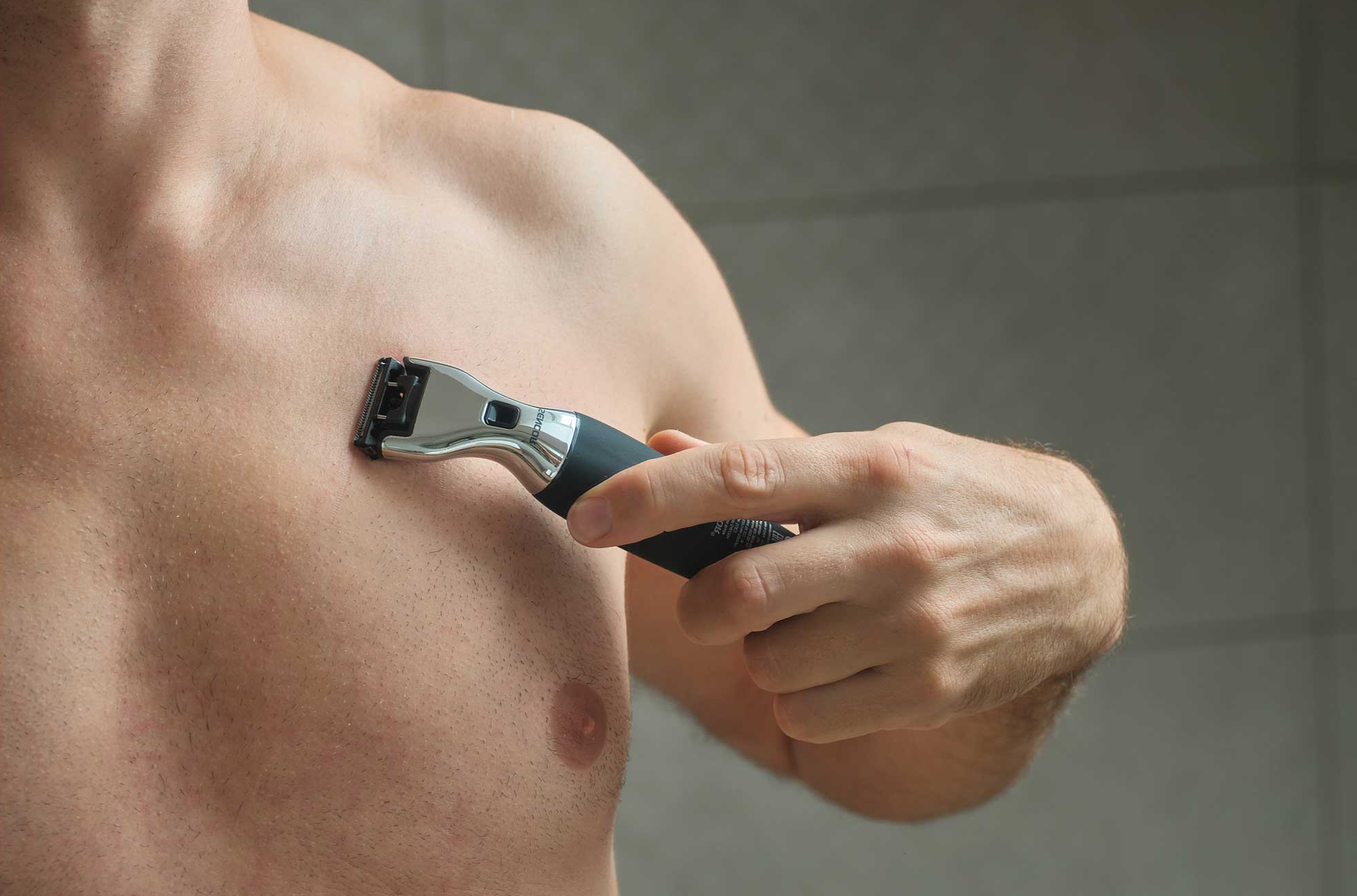 Egy férfi elektromos borotvával nyírja mellkasát szürke háttér előtt.