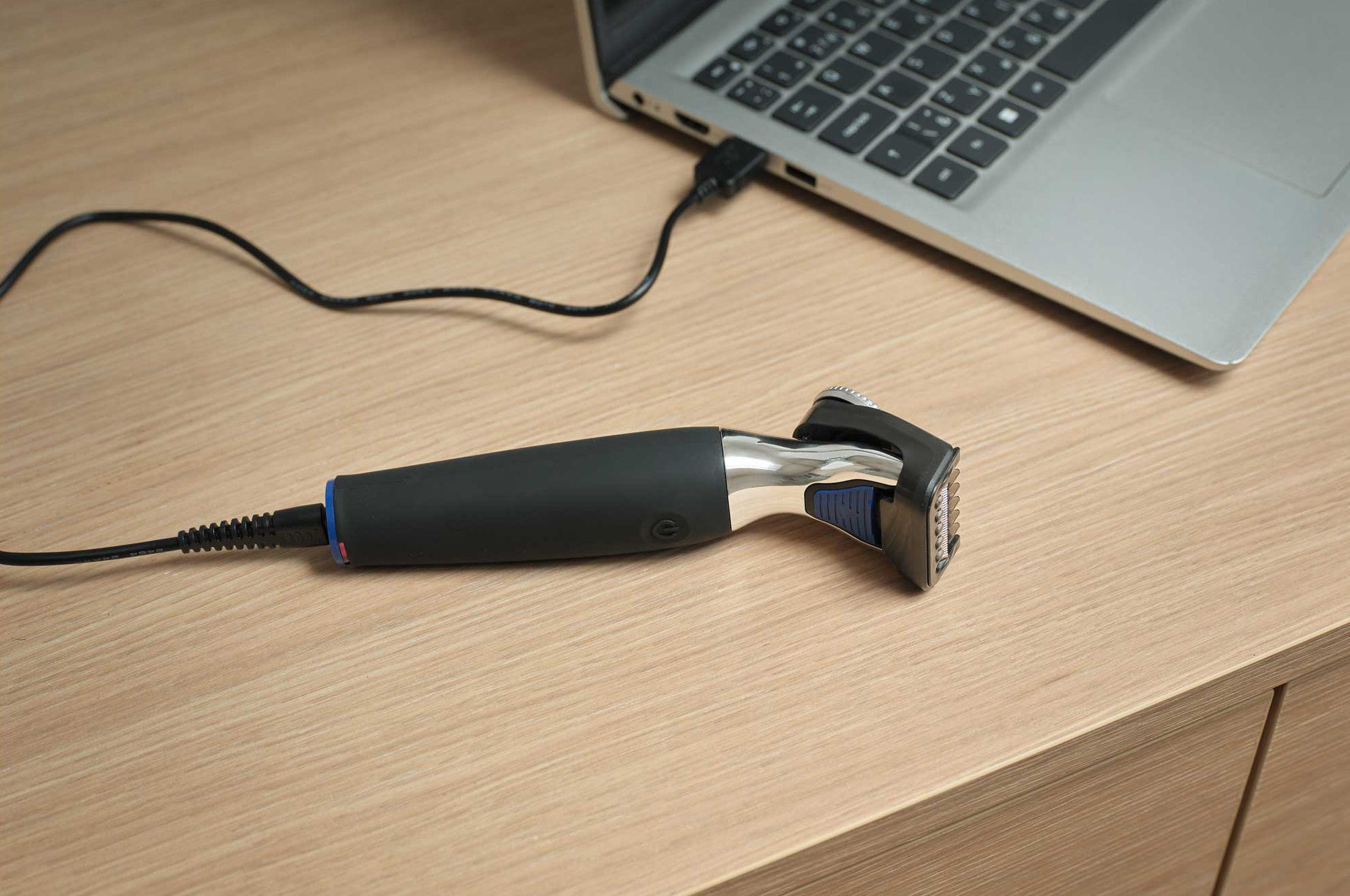 Egy fekete, króm részletekkel ellátott elektromos borotva USB-kábelen keresztül csatlakozik egy laptophoz.