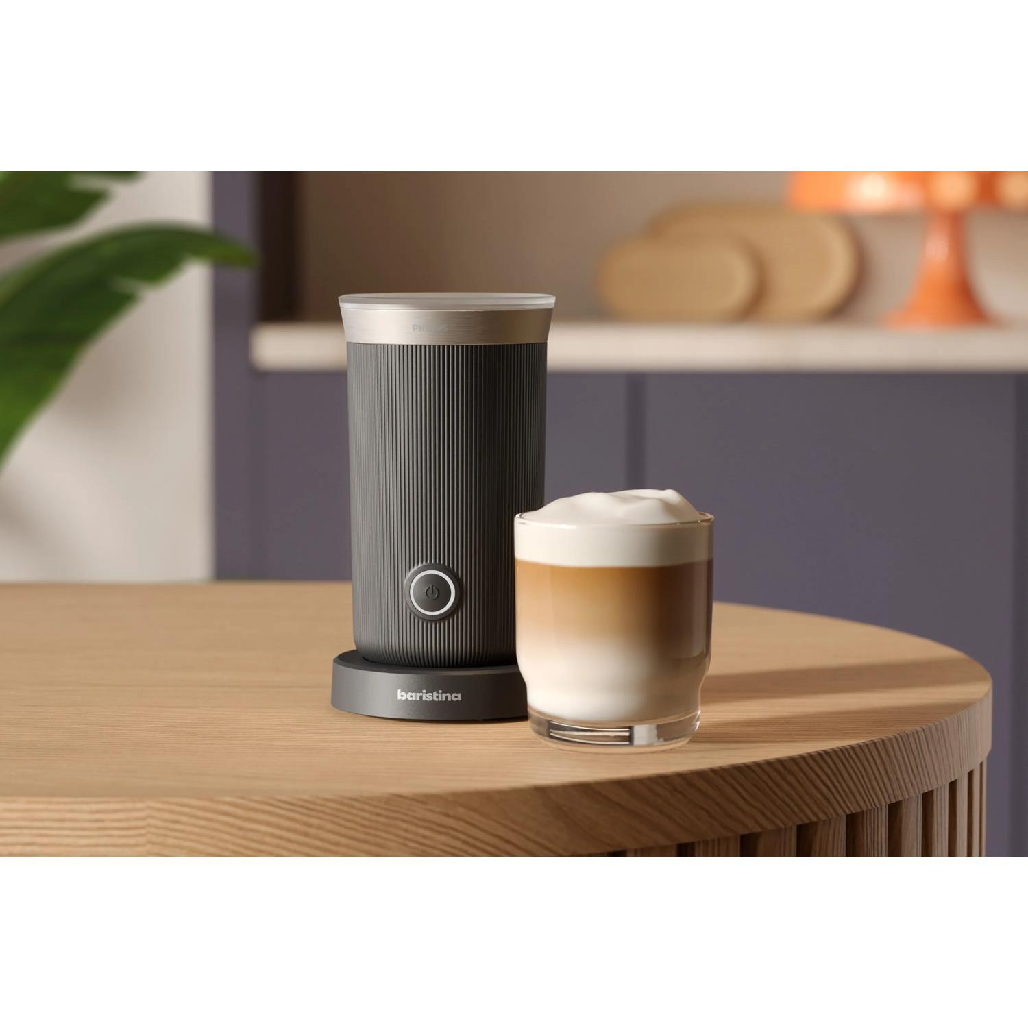 Un espumador de leche oscuro junto a un vaso de latte macchiato sobre una mesa de madera.