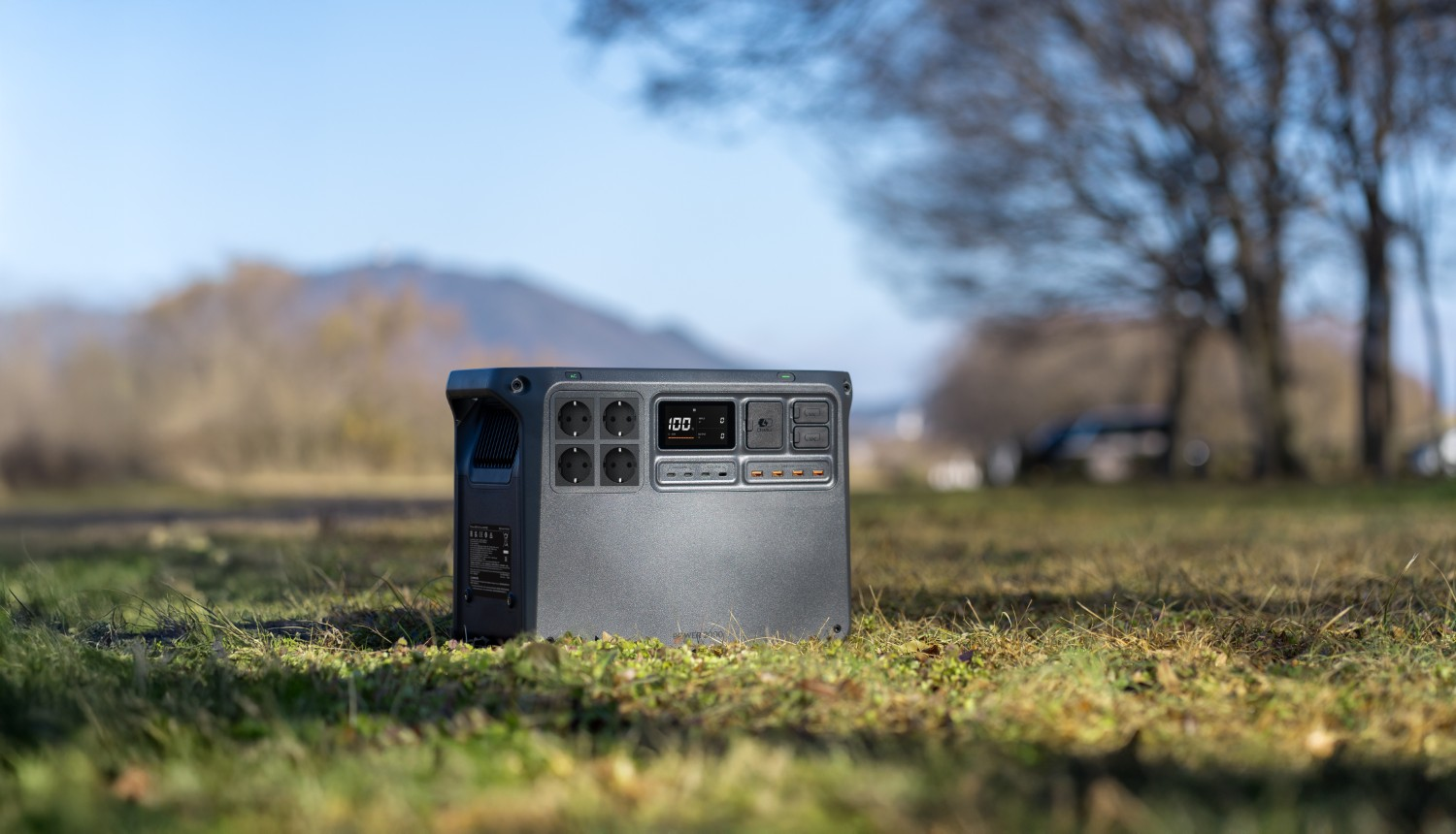 Szara elektrownia stoi na trawie. W tle drzewa i wzgórza.