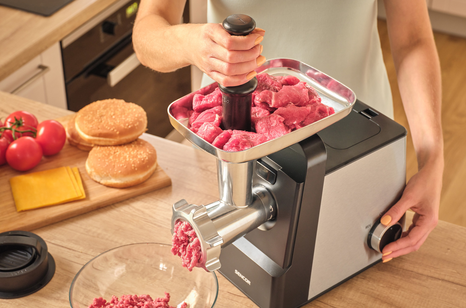 Egy személy darálót használ nyers hús feldolgozására. Burger zsemlék és paradicsomok vannak a közelben.