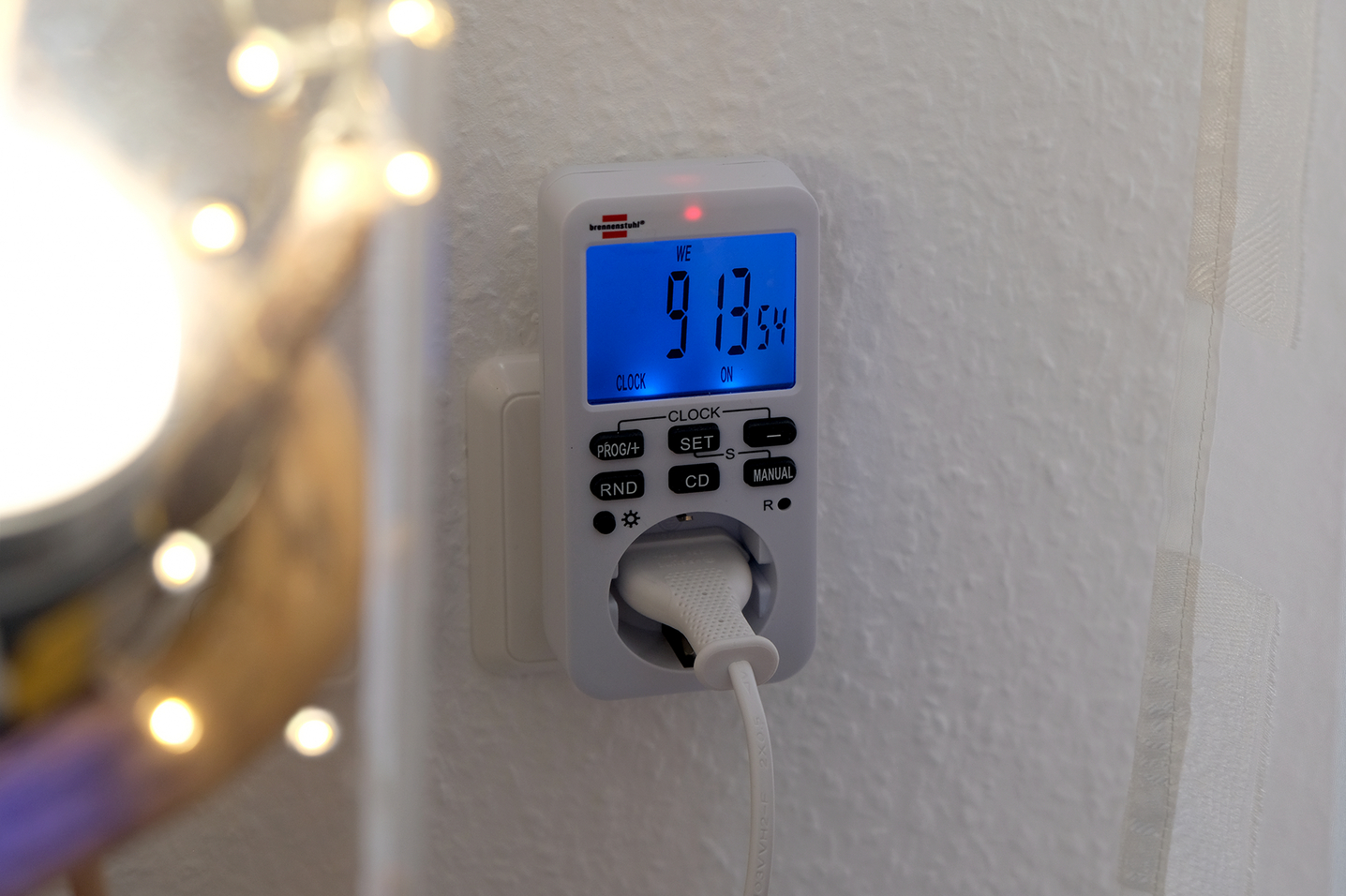 Ein digitaler Stromzähler mit blauem Display und Tasten an einer weißen Wand.