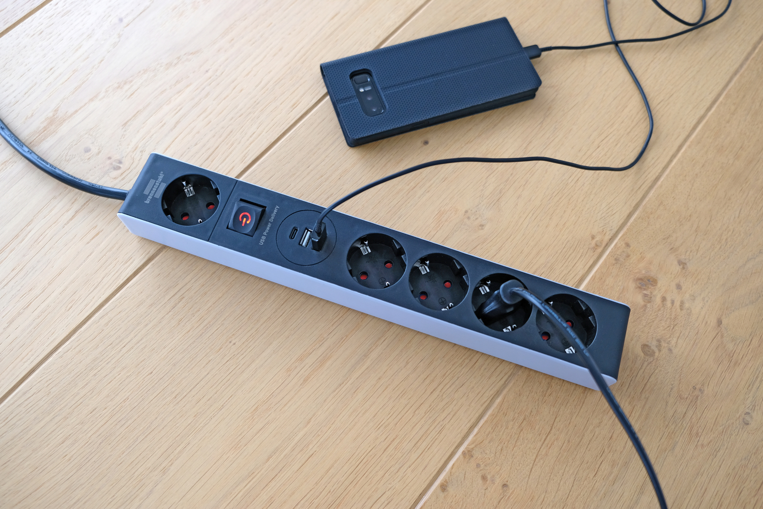 Schwarze Steckdosenleiste mit USB-Anschlüssen, die an ein Smartphone und ein Kabel angeschlossen sind.