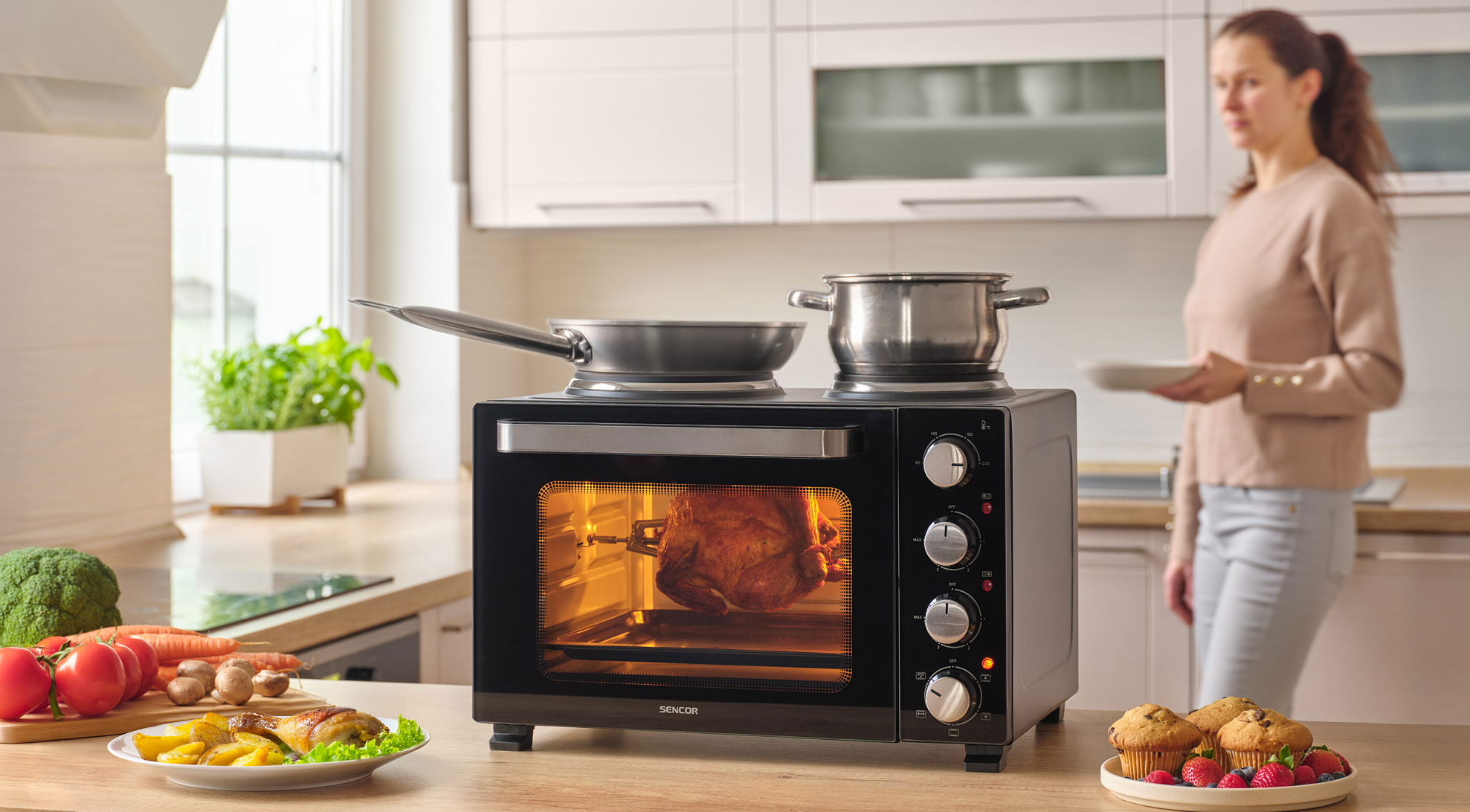 Egy rotisserie csirke forog a sütőben. Egy fazék és egy serpenyő áll mellette.