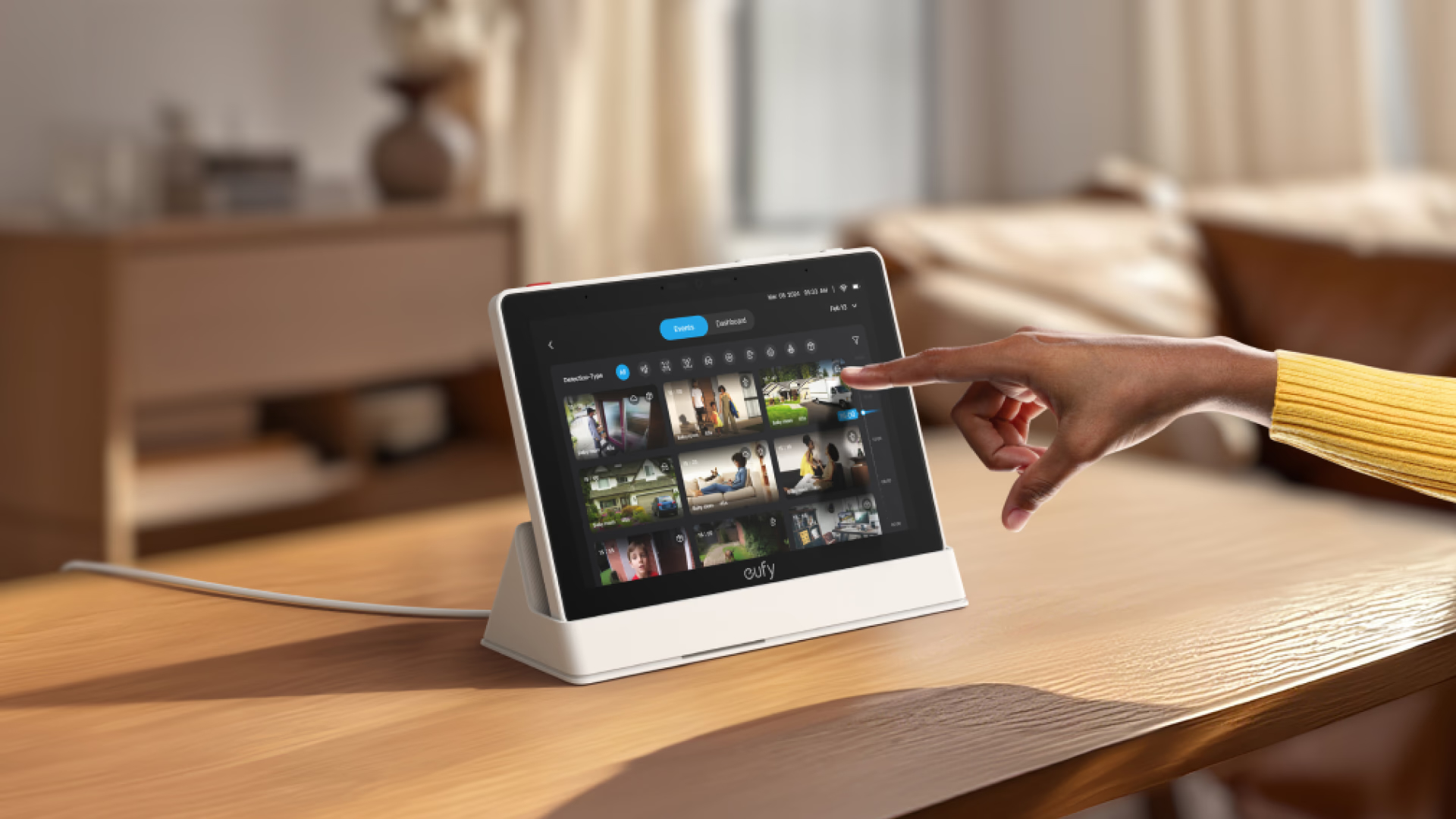 Eine Hand tippt auf einen Tablet-Bildschirm mit Video-Feeds. Ein Tisch und ein Hintergrund sind sichtbar.