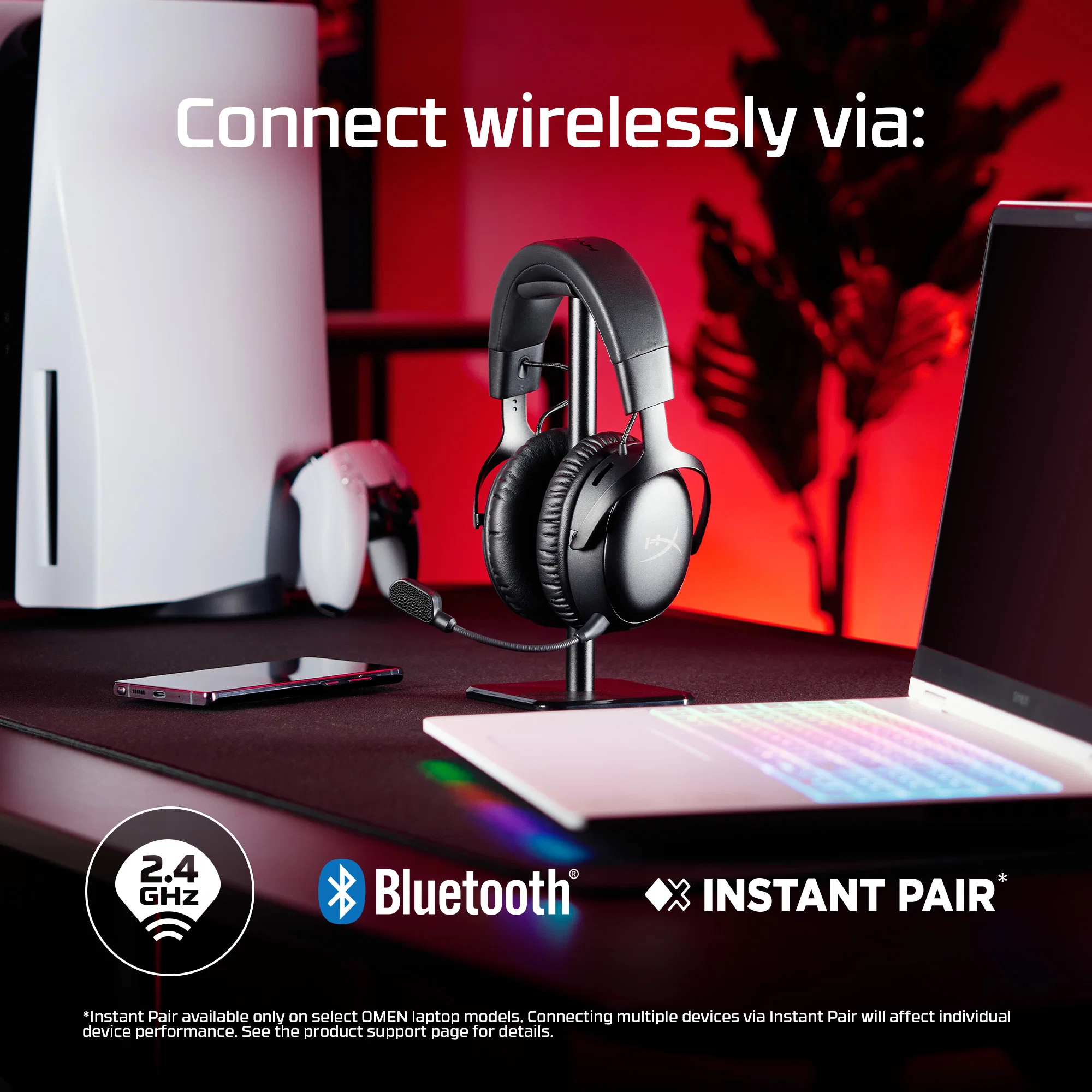 Cuffie su supporto con console, controller, telefono e laptop sullo sfondo.