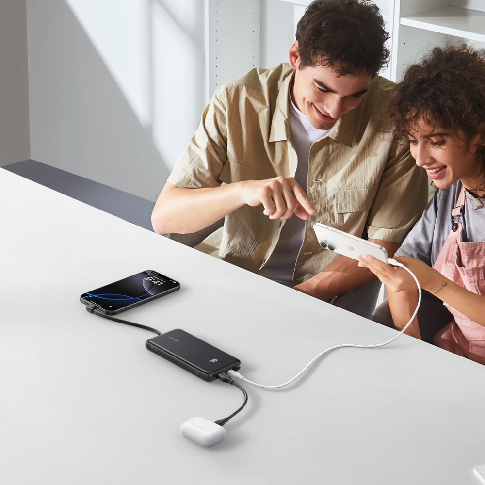 Mann und Frau laden Handys und Ohrhörer mit einer Powerbank auf einem weißen Tisch.