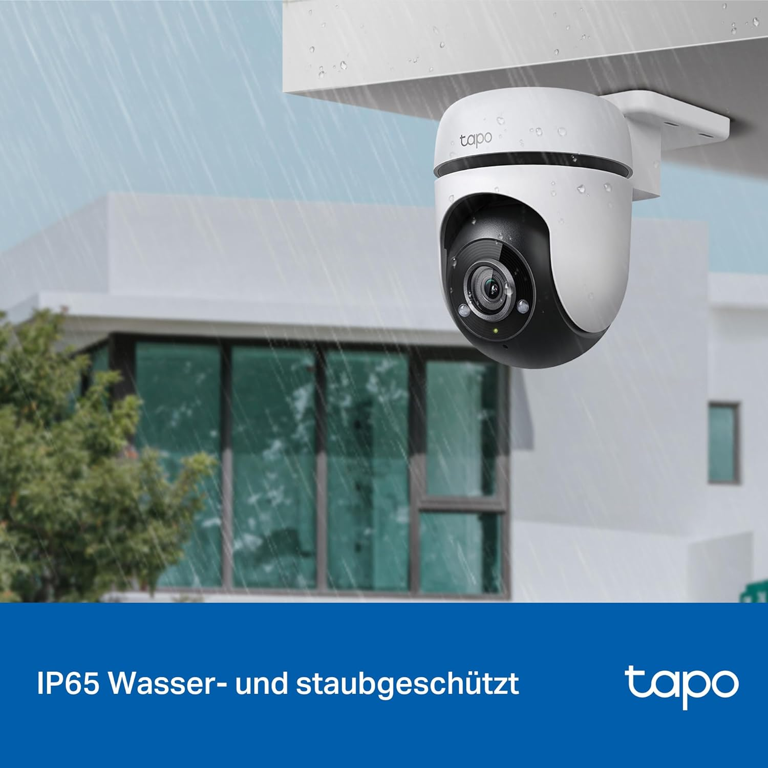 Eine weiße Überwachungskamera hängt unter einem Dach bei Regen.