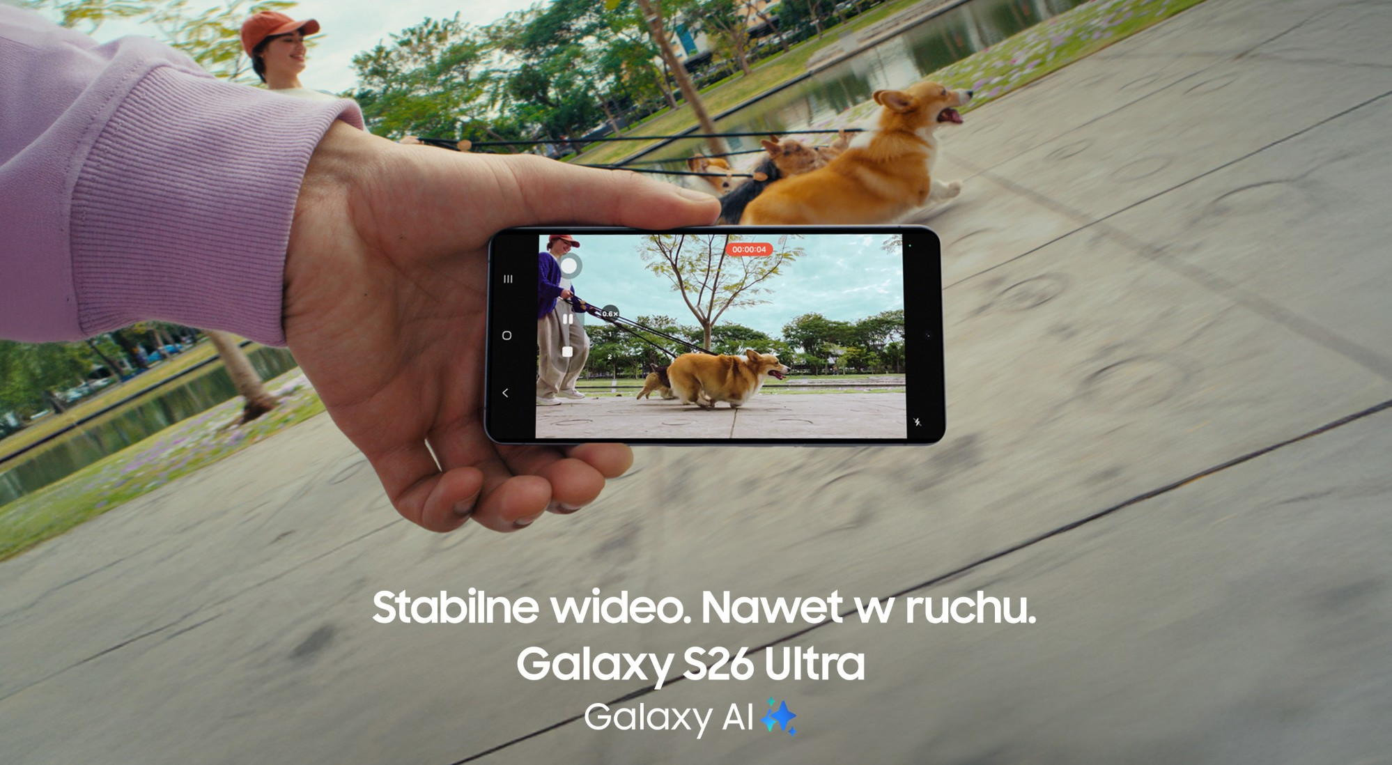 Dłoń trzyma smartfon pokazujący mężczyznę spacerującego z corgi w parku.