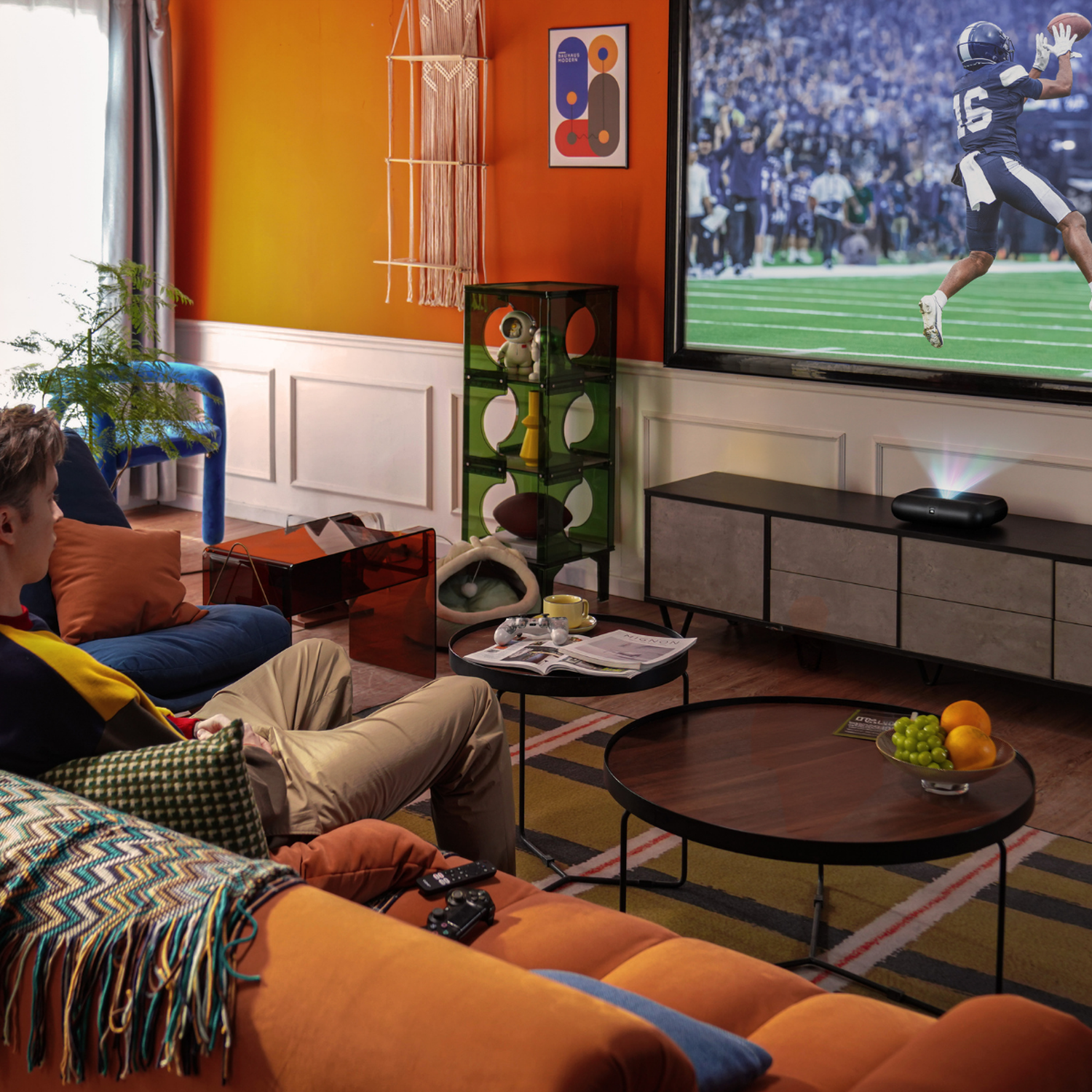 Man watches football on a large screen TV in a living room with orange walls and modern furniture.