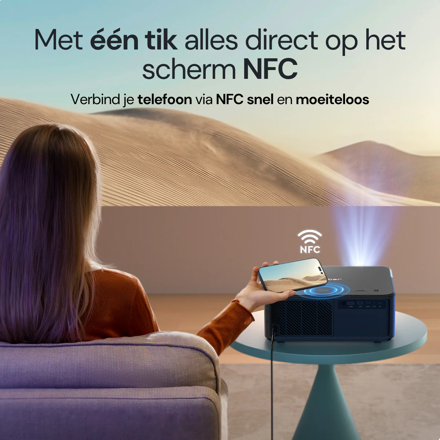 Person holds phone over projector displaying desert scene. NFC symbol above device.