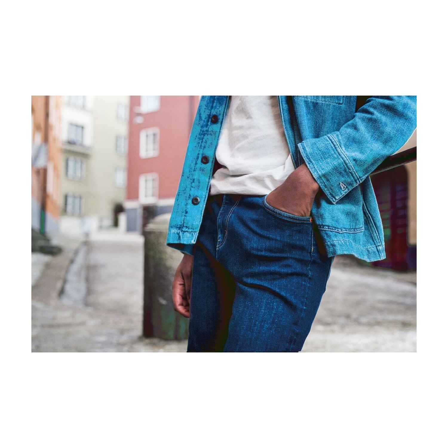 Uomo in jeans e giacca blu in un vicolo con edifici sullo sfondo.