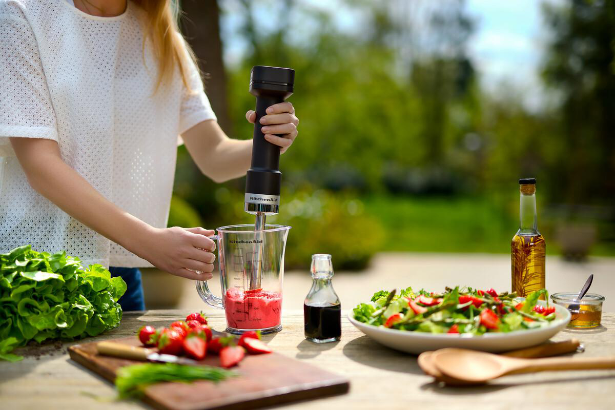 Eine Person mixt einen roten Smoothie im Freien mit Salat, Erdbeeren und Öl.