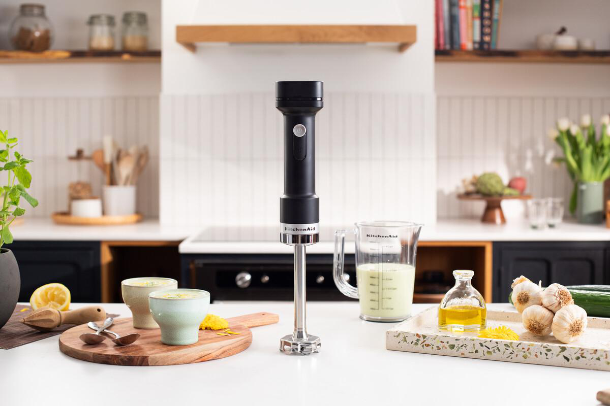 Ein schwarzer Stabmixer steht auf einer Arbeitsplatte neben Bechern, Öl, Knoblauch und Gemüse.