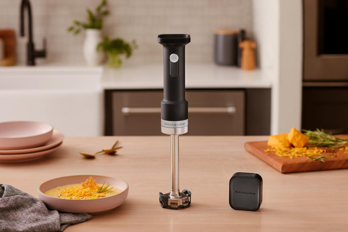 Ein Handmixer mit Zubehör steht auf einem Holztisch, daneben eine Schüssel Suppe.