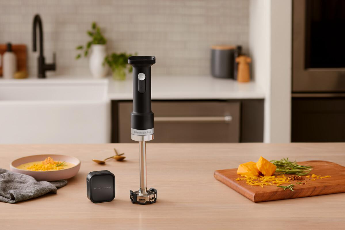 Ein schwarzer Stabmixer steht auf einem Holztisch neben einer Schale Suppe und Käse.