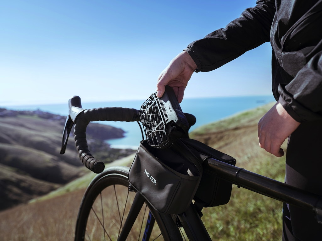 Eine Person legt ein Gerät auf eine Fahrradtasche, mit Meer und Hügeln im Hintergrund.