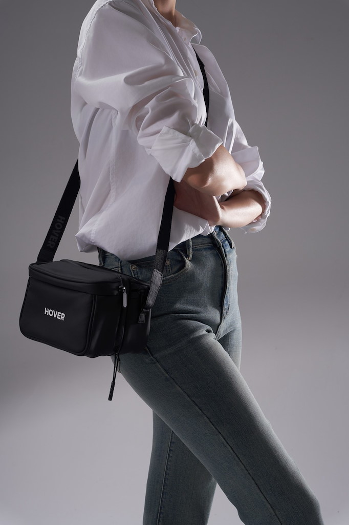 Eine Person mit weißem Hemd und Jeans trägt eine schwarze Umhängetasche mit der Aufschrift HOVER.