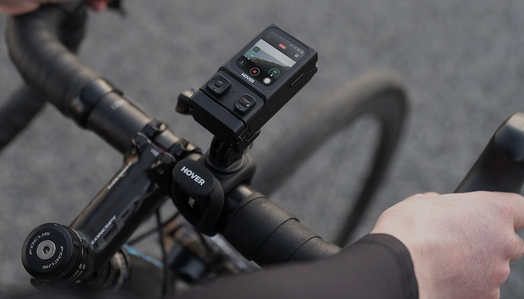 Eine schwarze Actionkamera ist am Fahrradlenker montiert. Hände einer Person halten die Griffe.