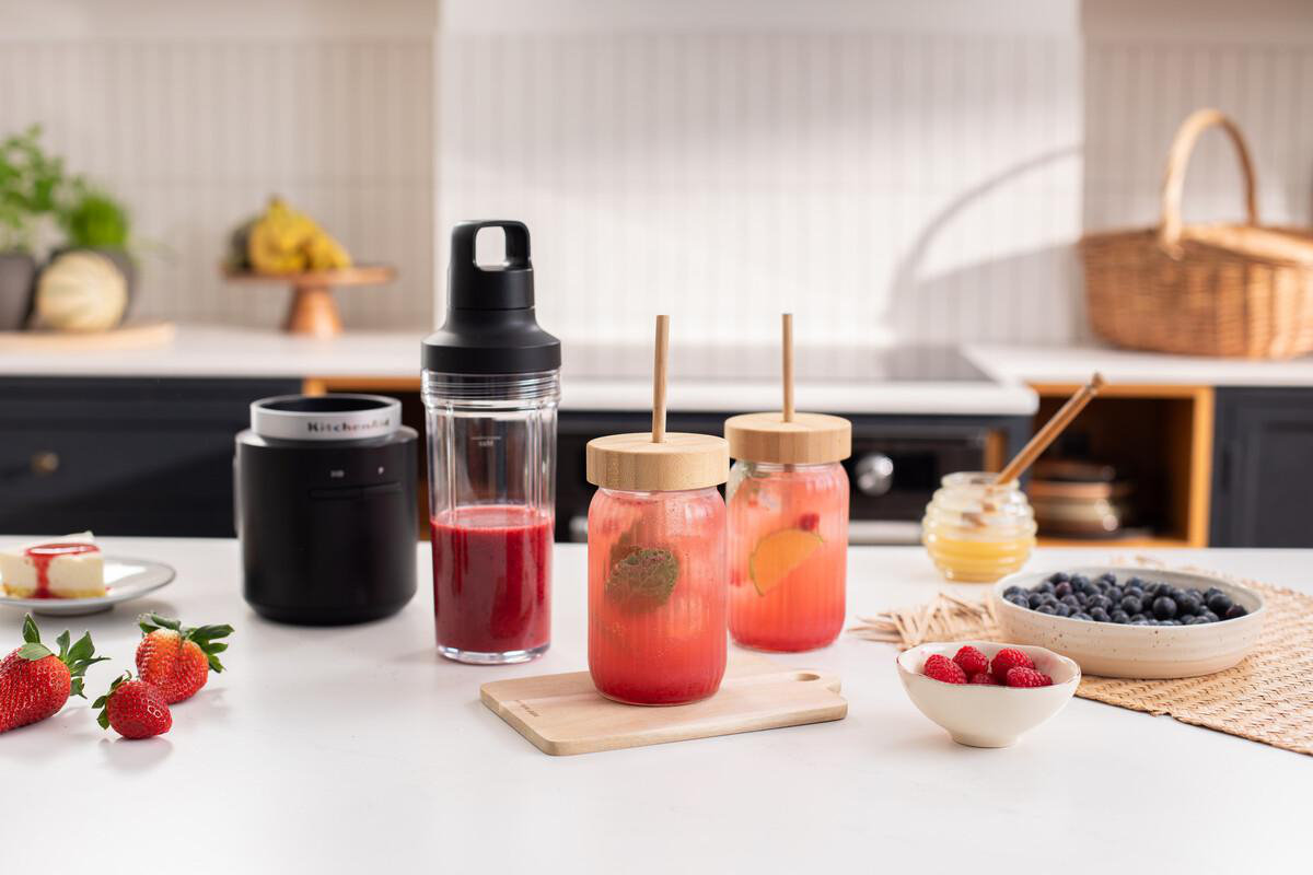 Ein Mixer, zwei Getränke, Beeren, Kuchen und Honig auf einer Küchentheke.