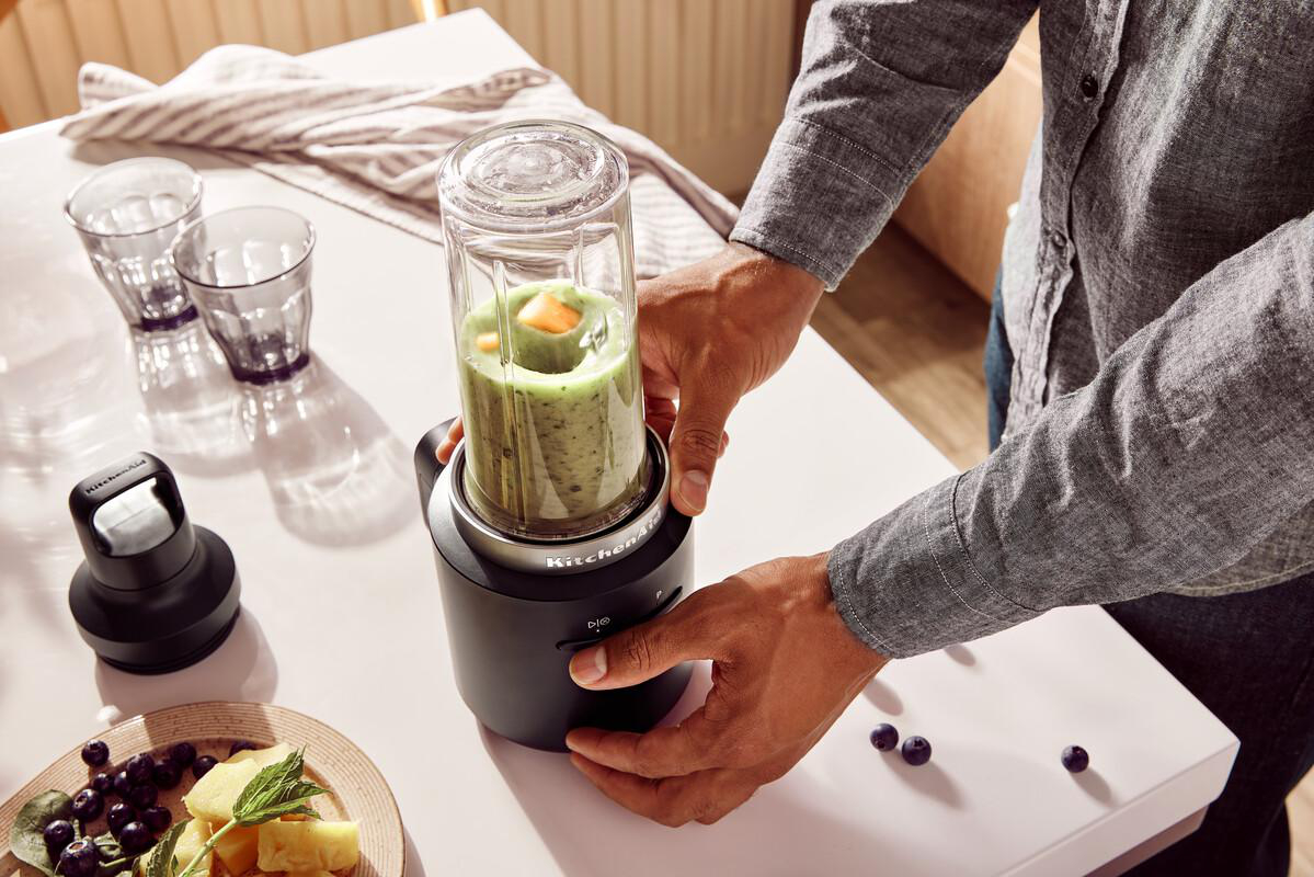 Ein Mann mixt einen grünen Smoothie in einem KitchenAid Mixer. Obst und Beeren sind in der Nähe.