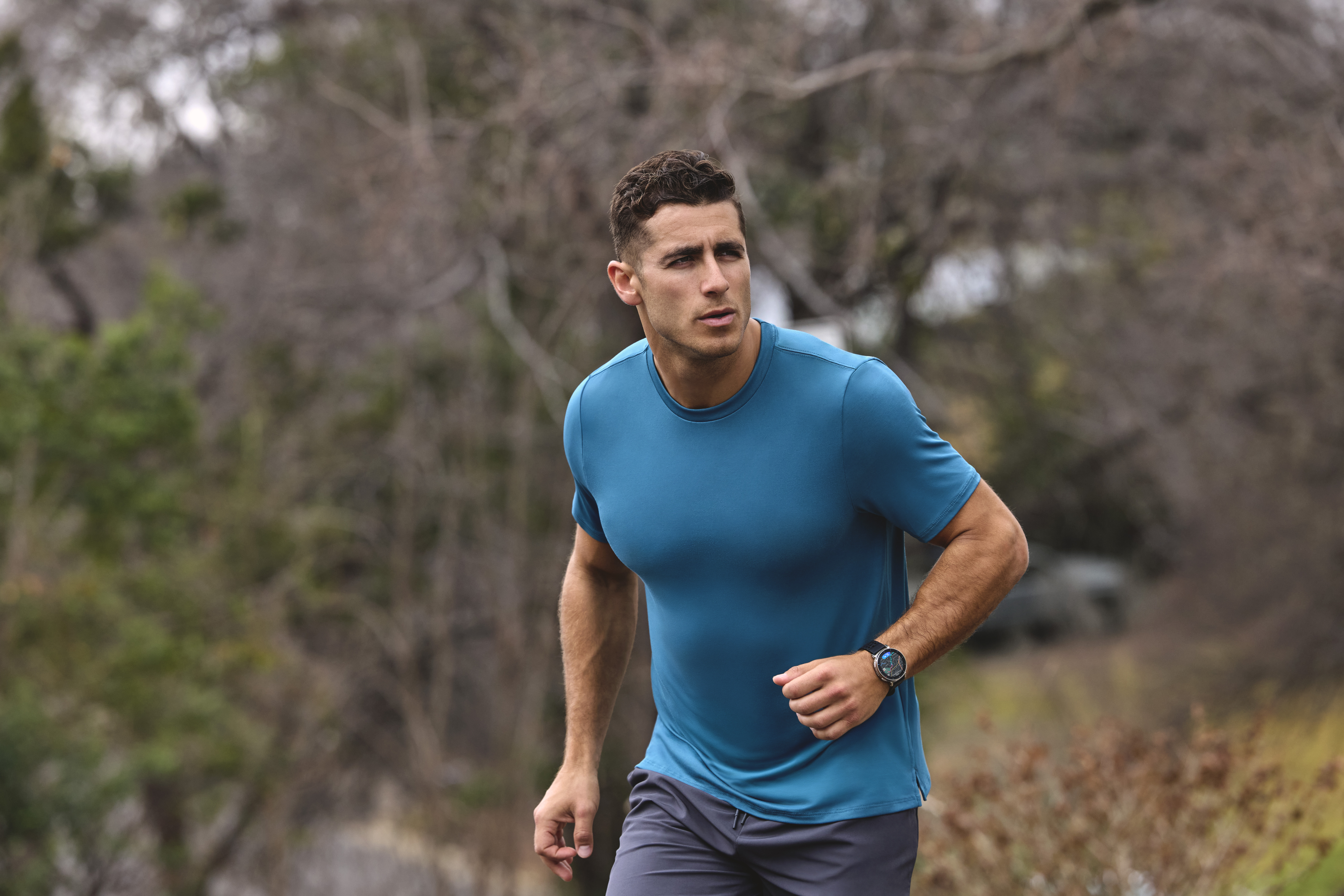 Ein Mann in einem blauen Shirt und grauen Shorts läuft im Freien mit Bäumen im Hintergrund.