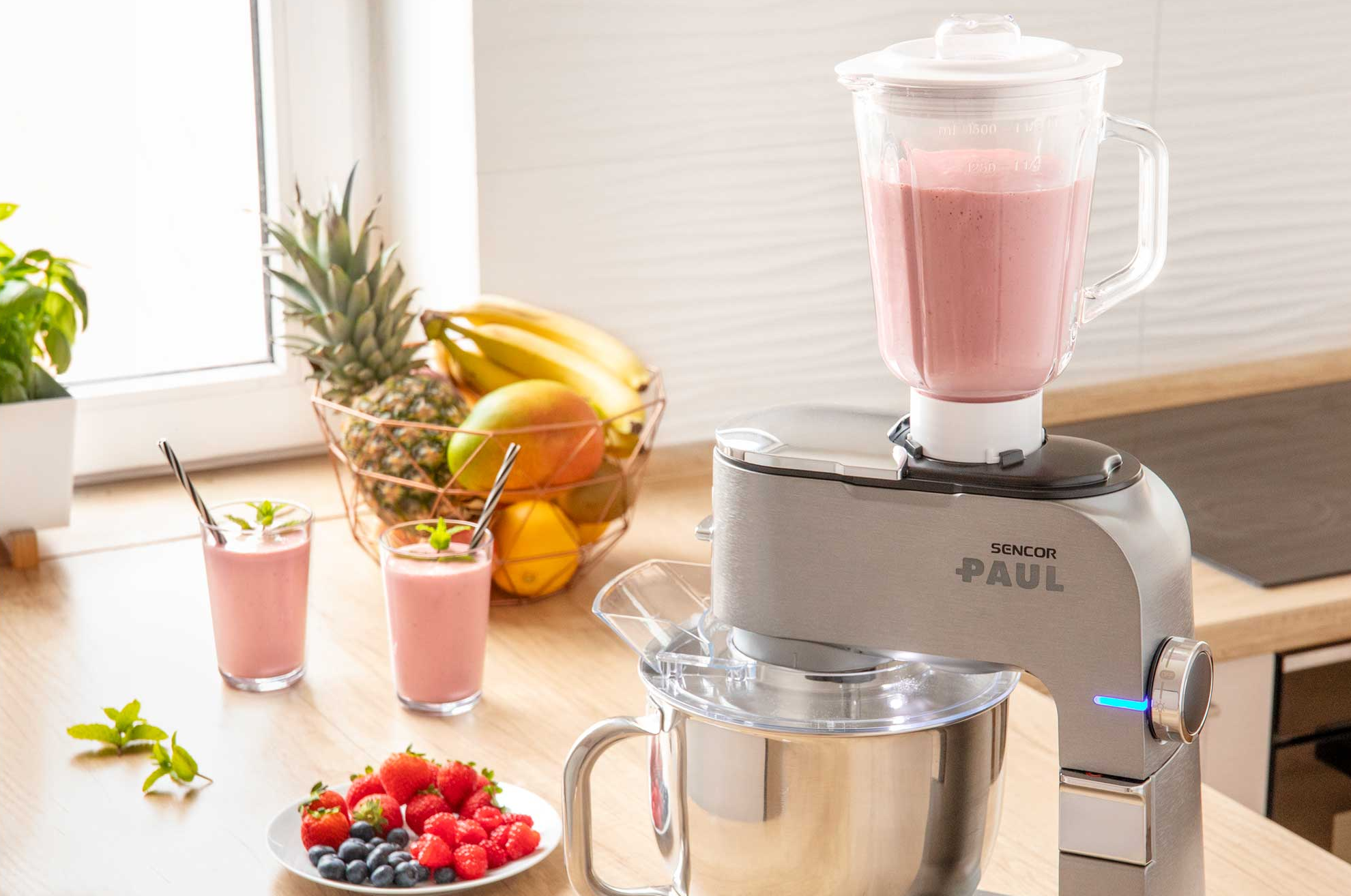 Ezüst konyhai robotgép rózsaszín smoothie-s pohárral és gyümölcsökkel.