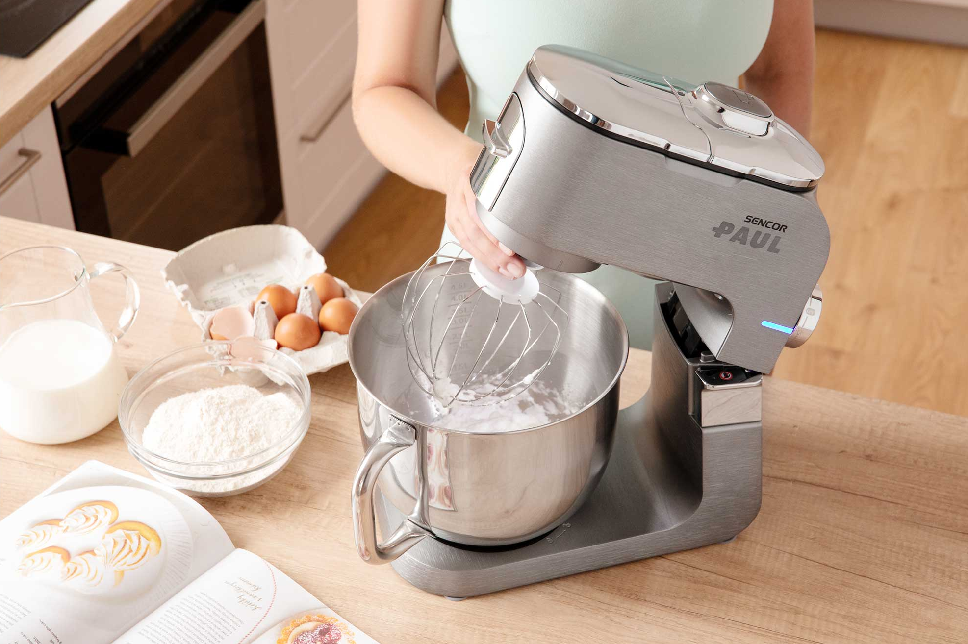 Egy álló mixer keverőtállal, tojásokkal, liszttel és tejjel faasztalon.
