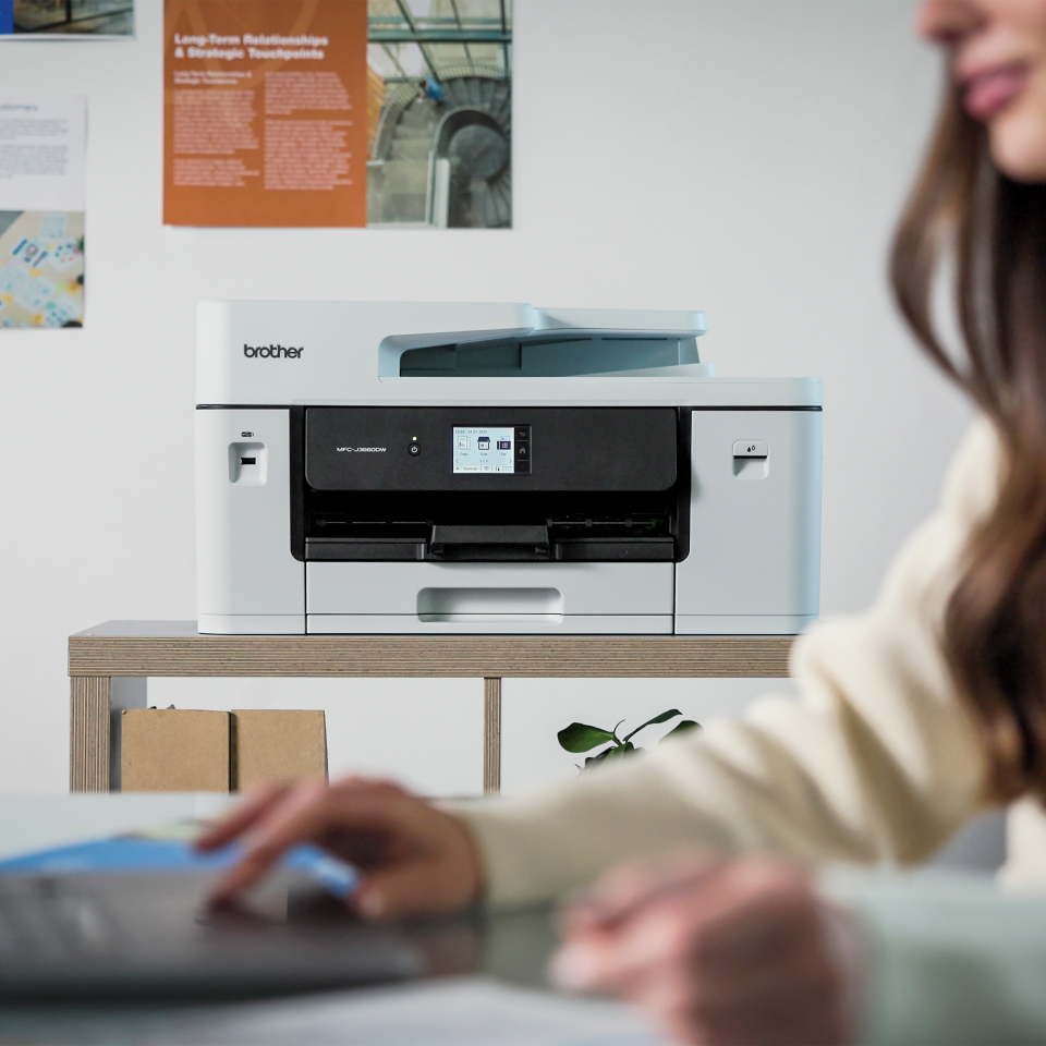 Biała drukarka Brother stoi na półce. Dłonie osoby są widoczne na laptopie na pierwszym planie.