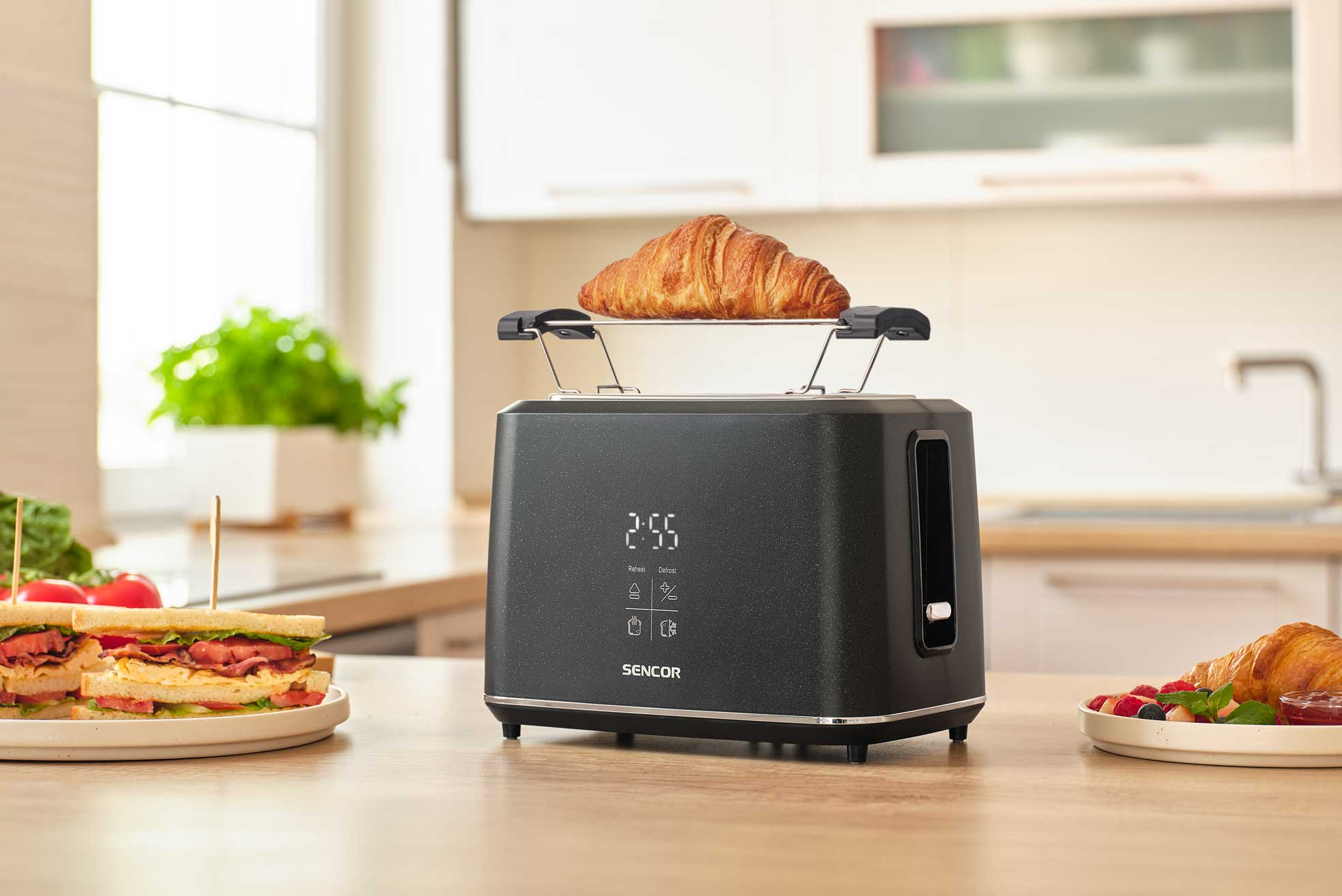 Fekete kenyérpirító croissanttal a rácsán. Közelben szendvicsek és gyümölcsök.