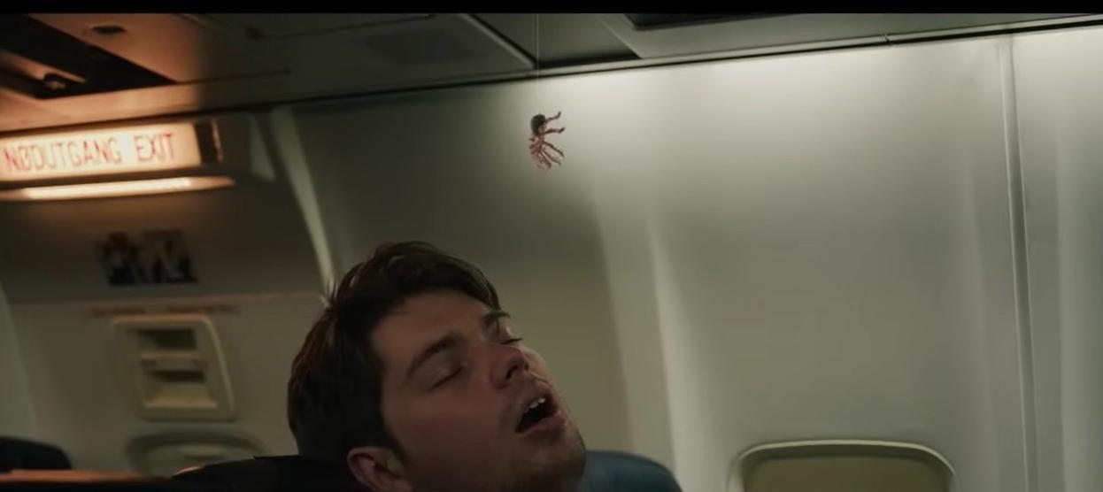 Ein Mann schläft im Flugzeug, während eine Spinne von oben herabhängt.