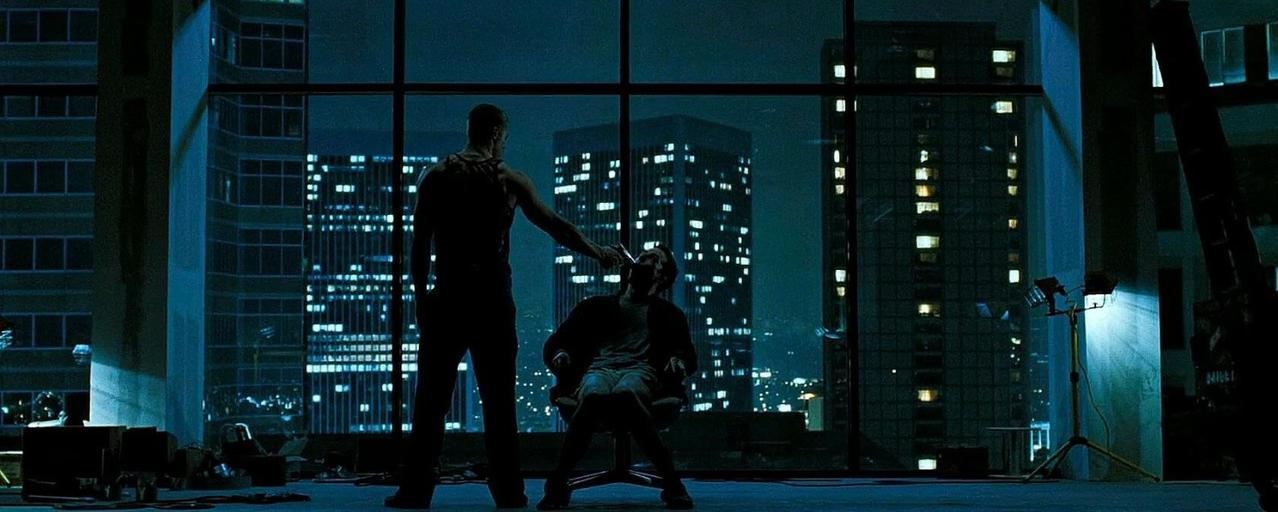 Zwei Männer stehen vor einem Fenster mit Blick auf eine Stadt bei Nacht. Einer hält ein Messer.