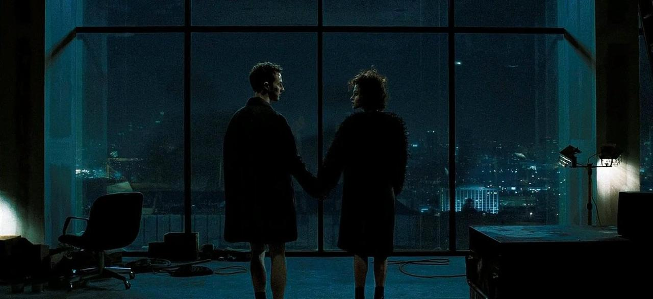 Zwei Menschen stehen vor einem Fenster mit Blick auf eine nächtliche Stadt.