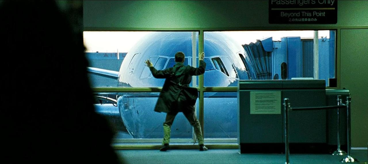 Mann in dunklem Mantel winkt einem Flugzeug mit ausgestreckten Armen zu.