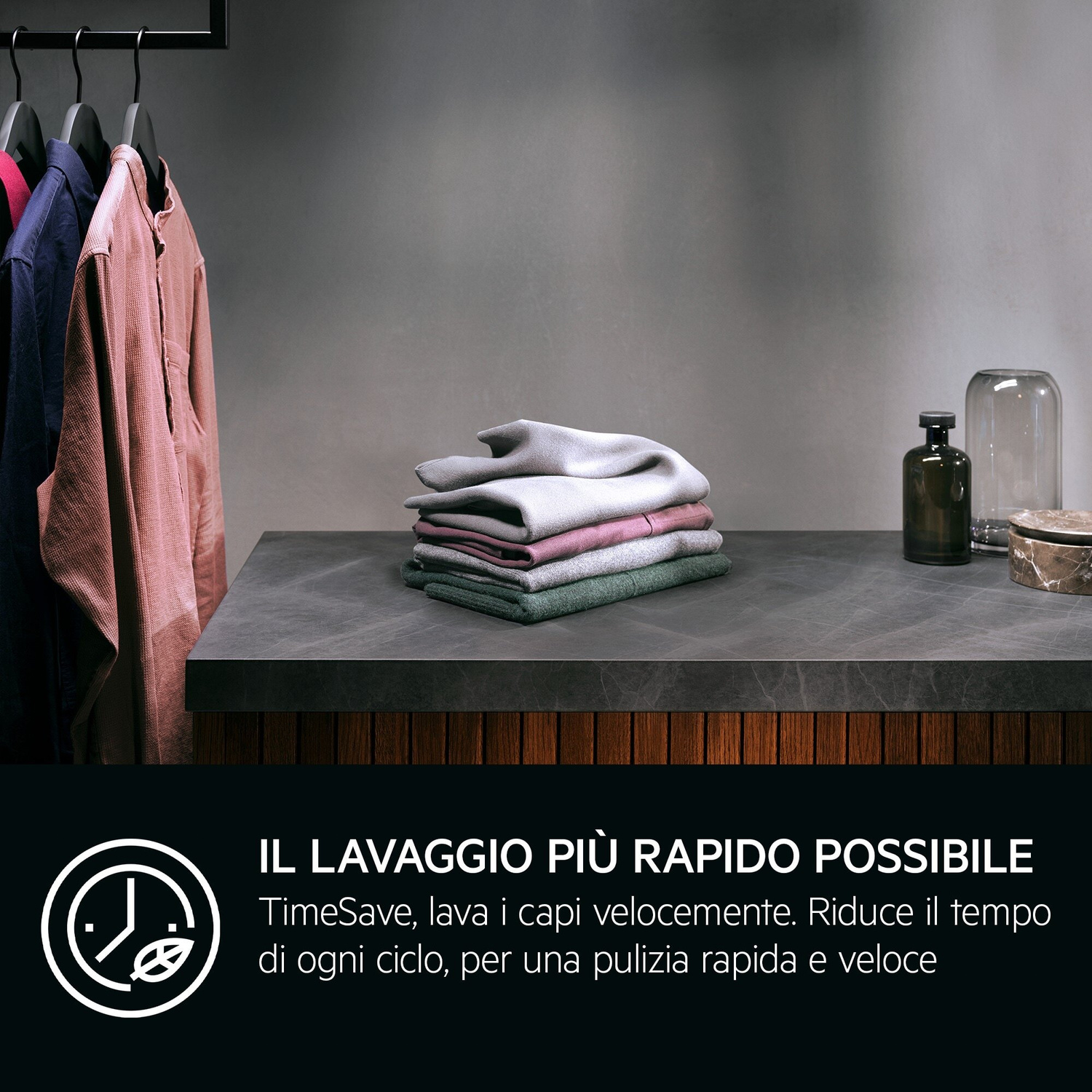 Vestiti appesi, tessuti impilati. Vasi di vetro e bottiglia su tavolo.