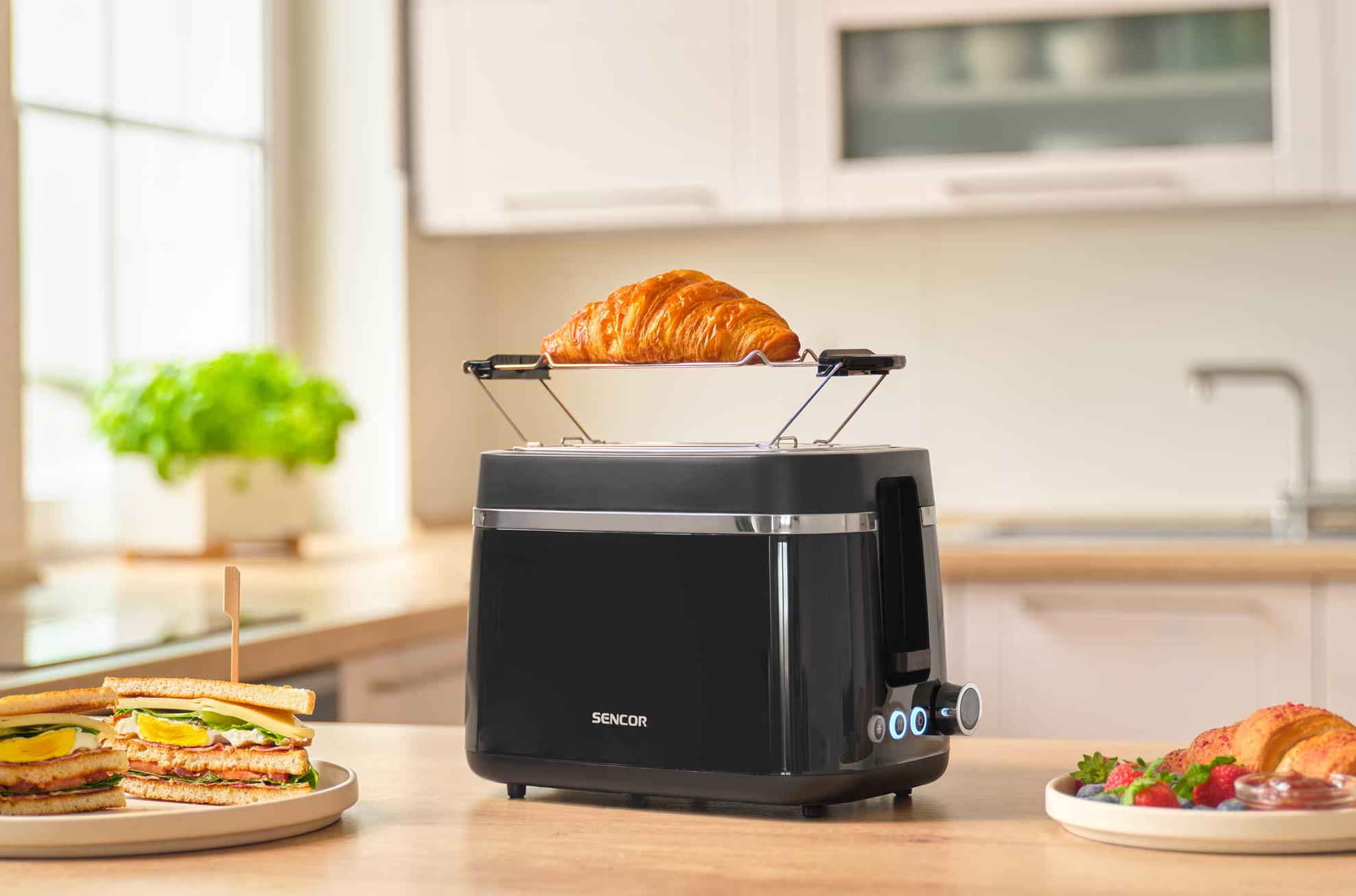 Egy fekete kenyérpirító, rajta croissant. Mellette szendvicsek és bogyós gyümölcsök.