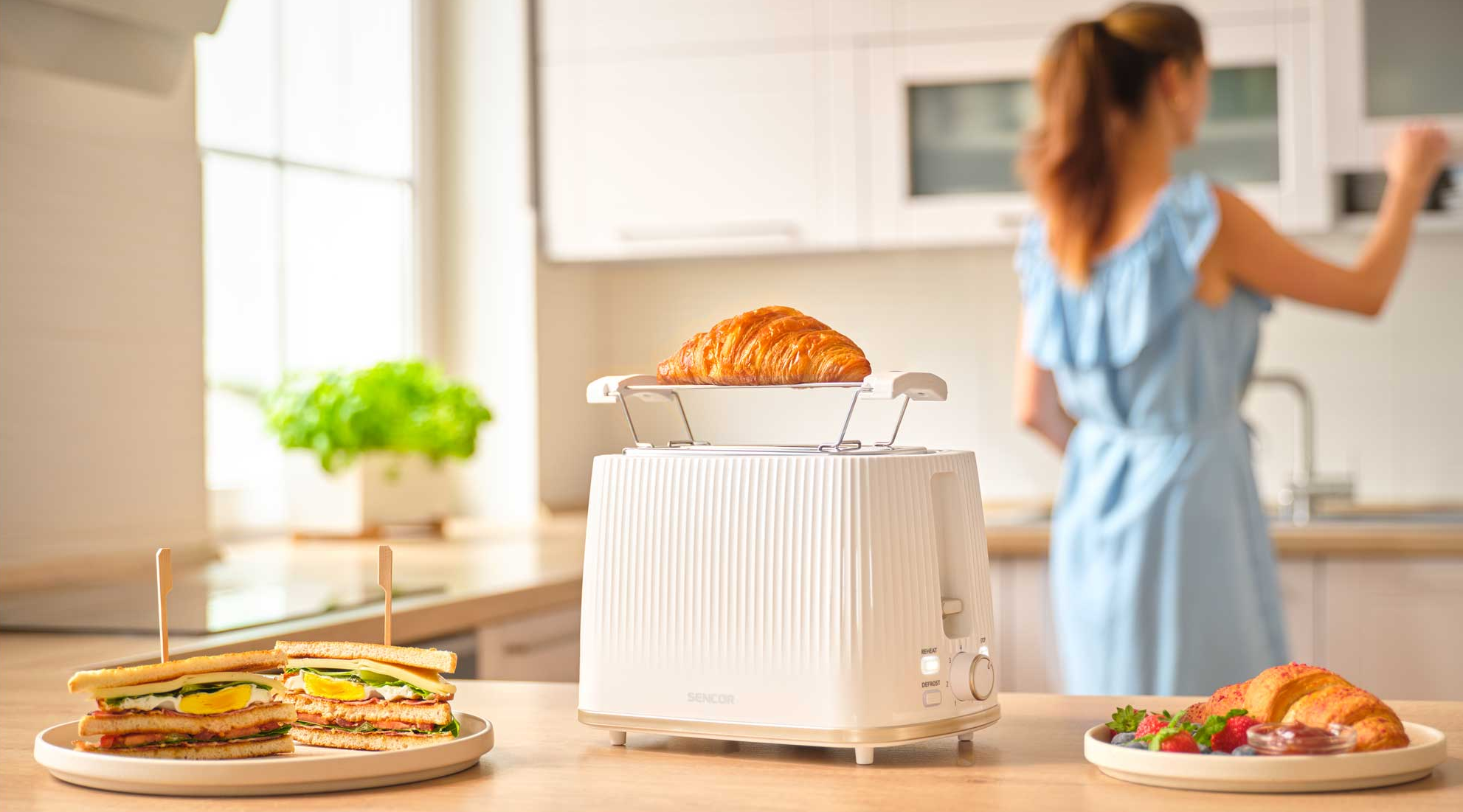 Egy fehér kenyérpirító croissanttal, szendvicsekkel és bogyós gyümölcsökkel faasztalon.