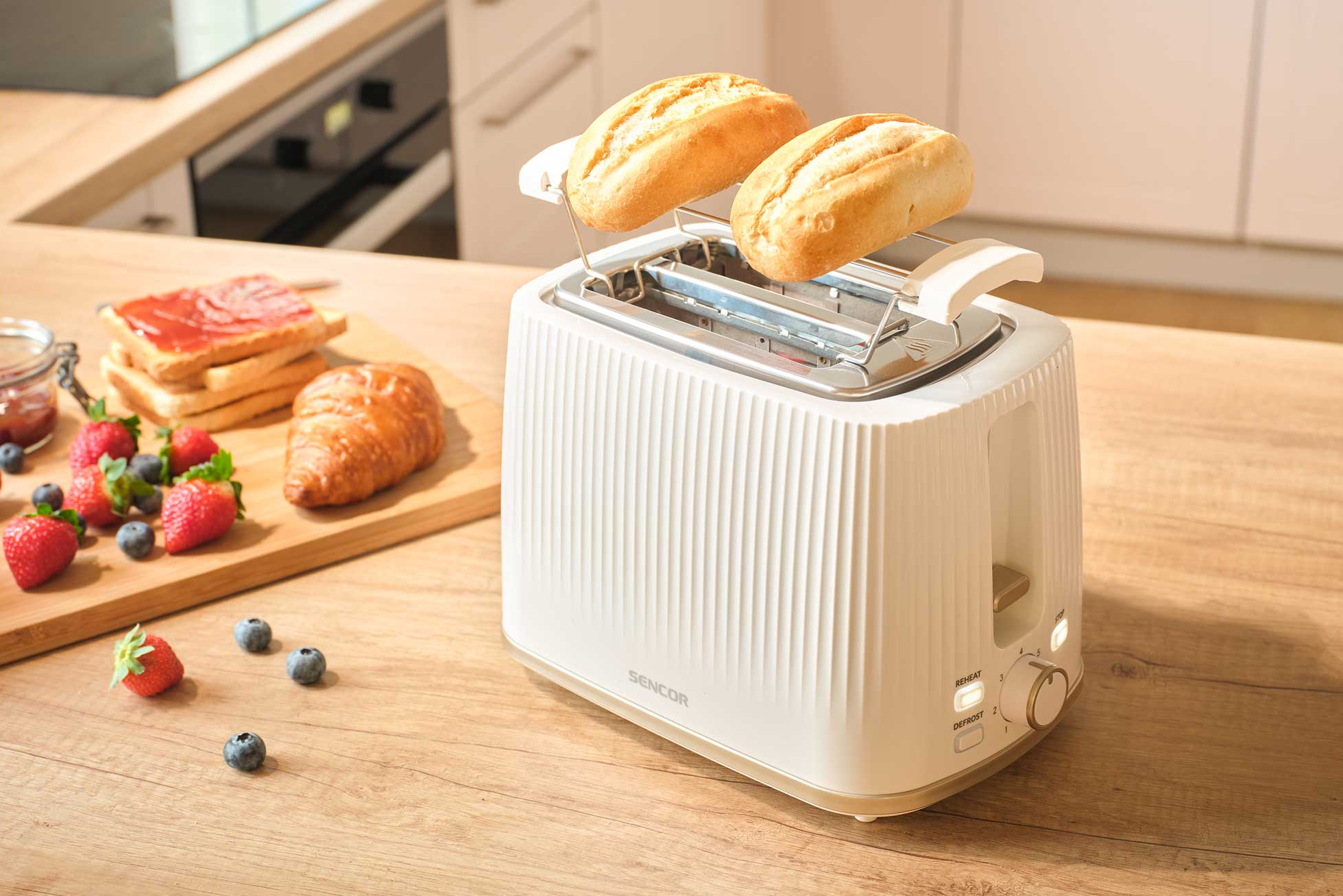 Fehér kenyérpirító két zsemlével, mellette pirítós, croissant és bogyók.