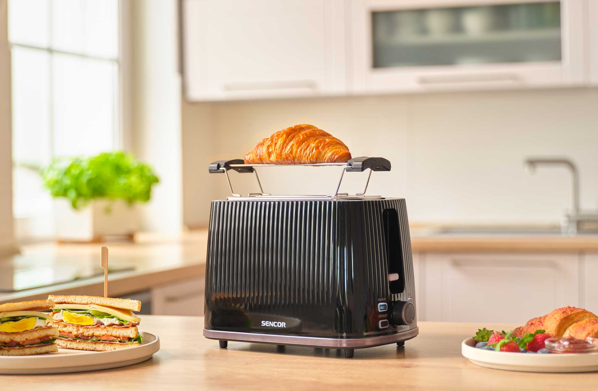 Egy fekete kenyérpirító croissanttal, szendvicsekkel és bogyós gyümölcsökkel faasztalon.