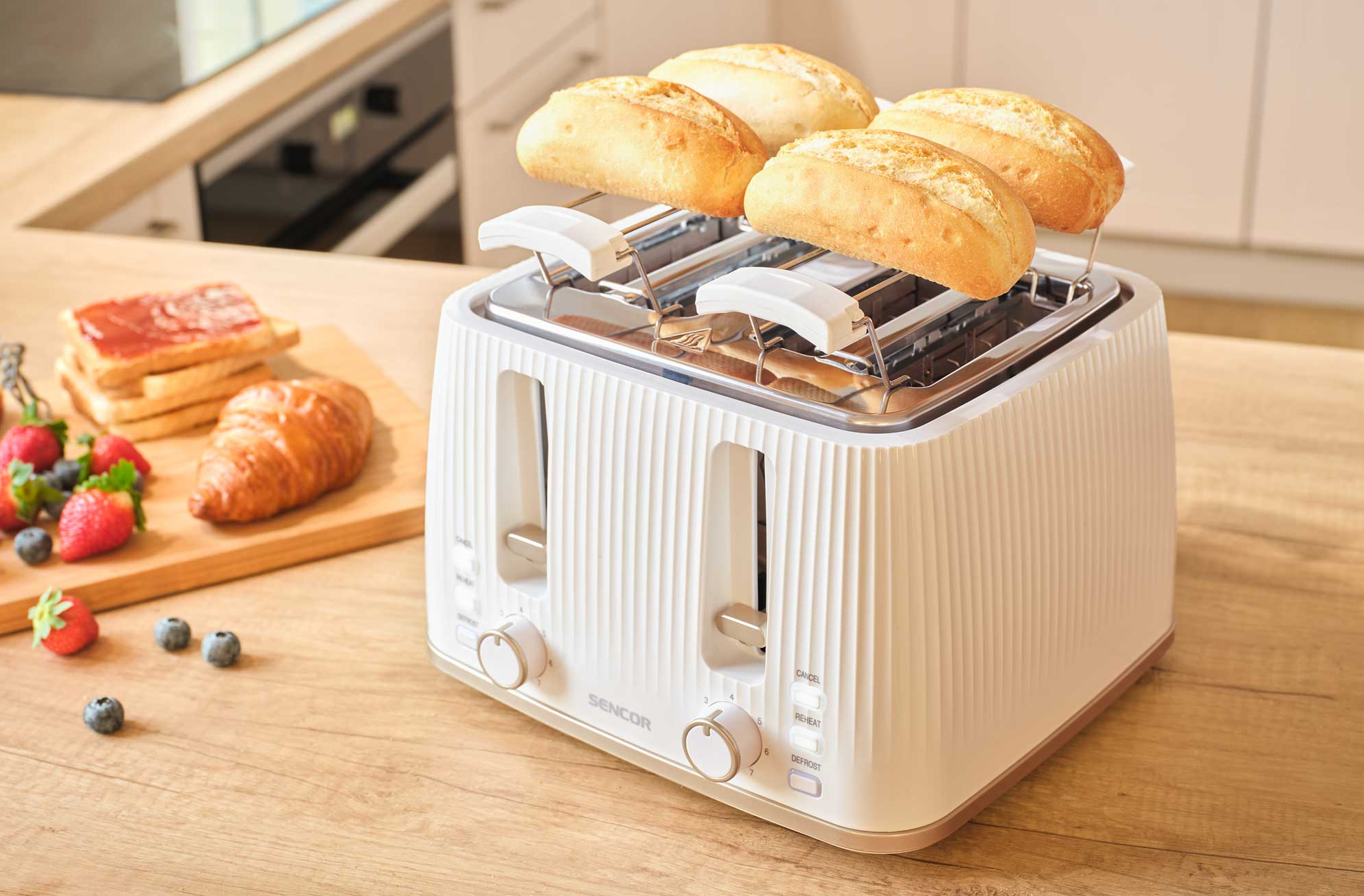 Fehér kenyérpirító négy zsemlével, kenyér, croissant és bogyók mellett egy faasztalon.
