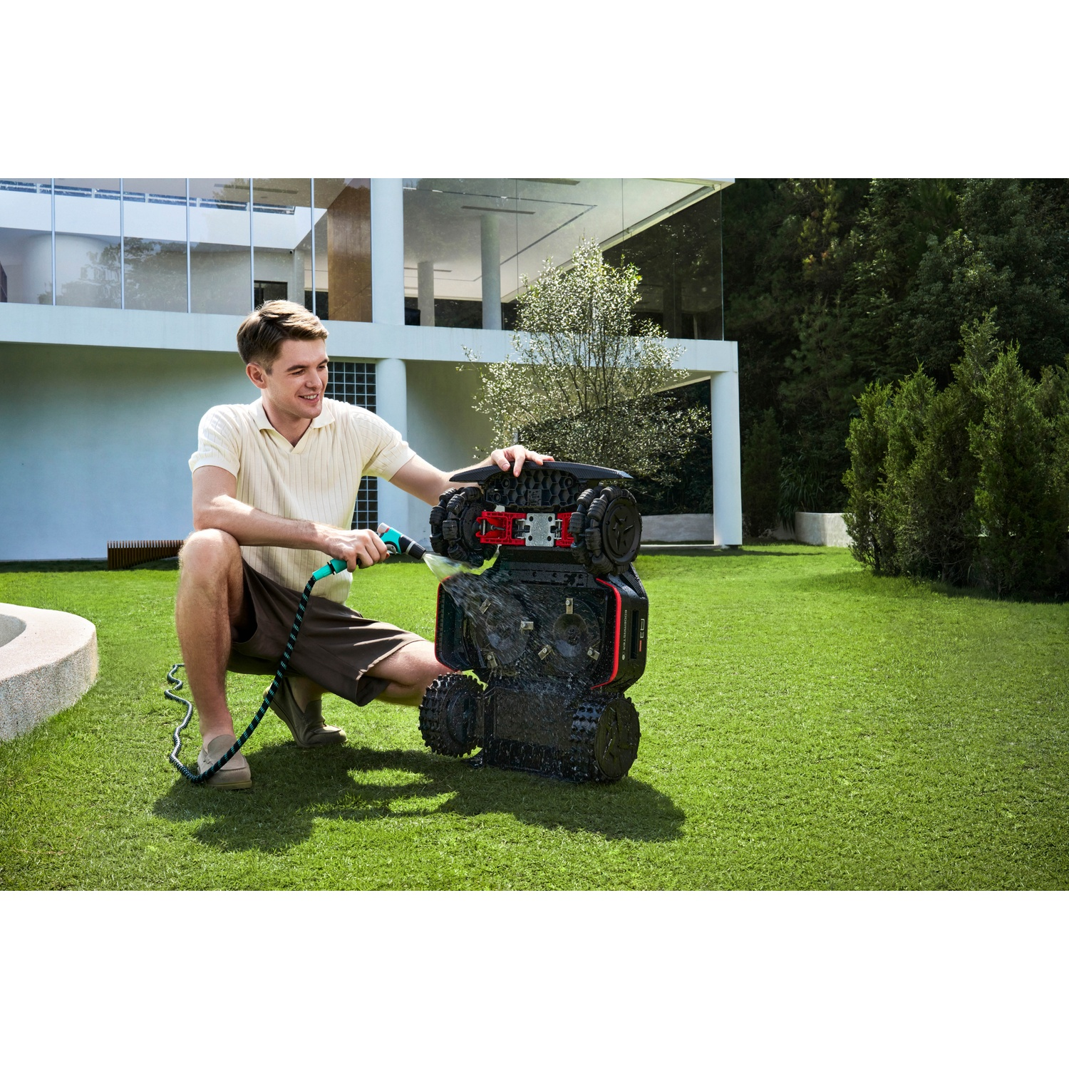 A man washes a lawnmower on a lawn in front of a modern house.